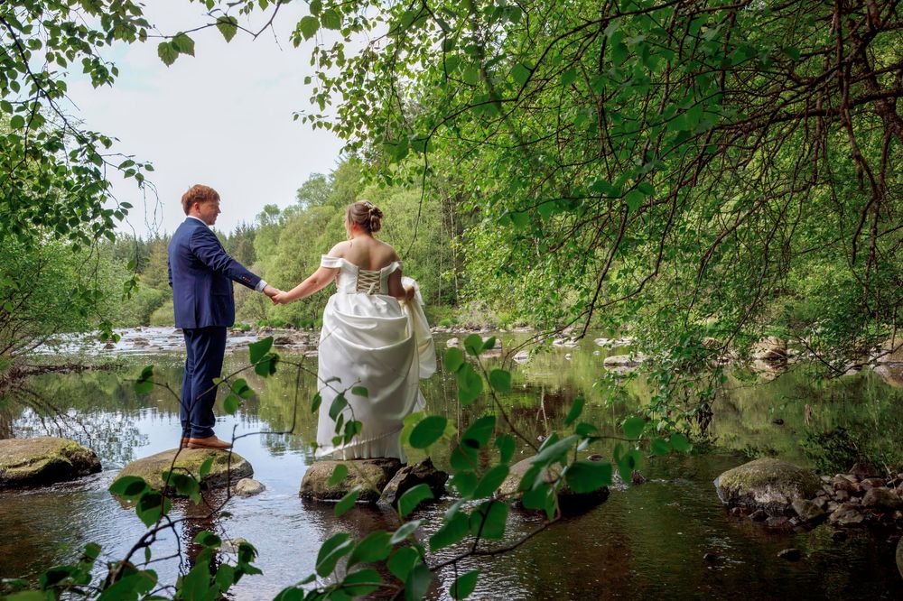 Bride and groom holding hands on river rocks surrounded by lush green trees on their wedding day.