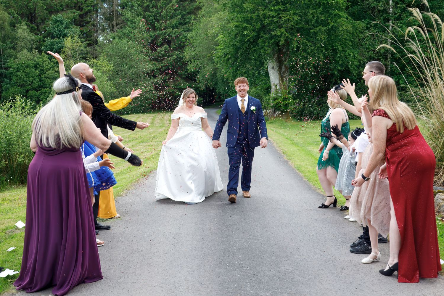 Bride and groom walk between cheering guests lining a path at outdoor wedding celebration.