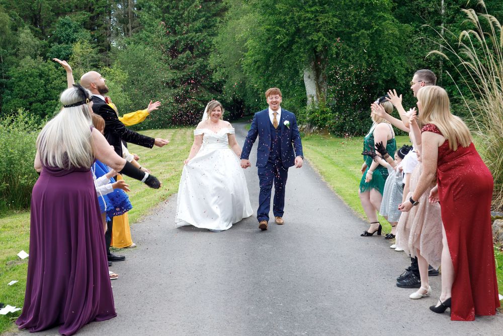 Bride and groom walk between cheering guests lining a path at outdoor wedding celebration.