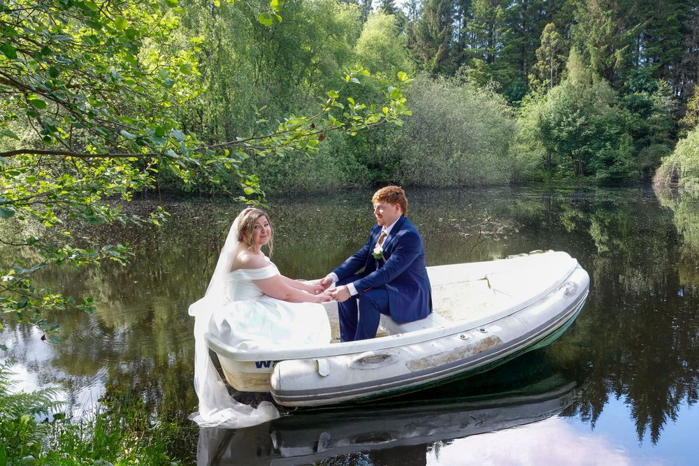Bride and groom sitting in a small boat on a calm lake surrounded by lush green trees.