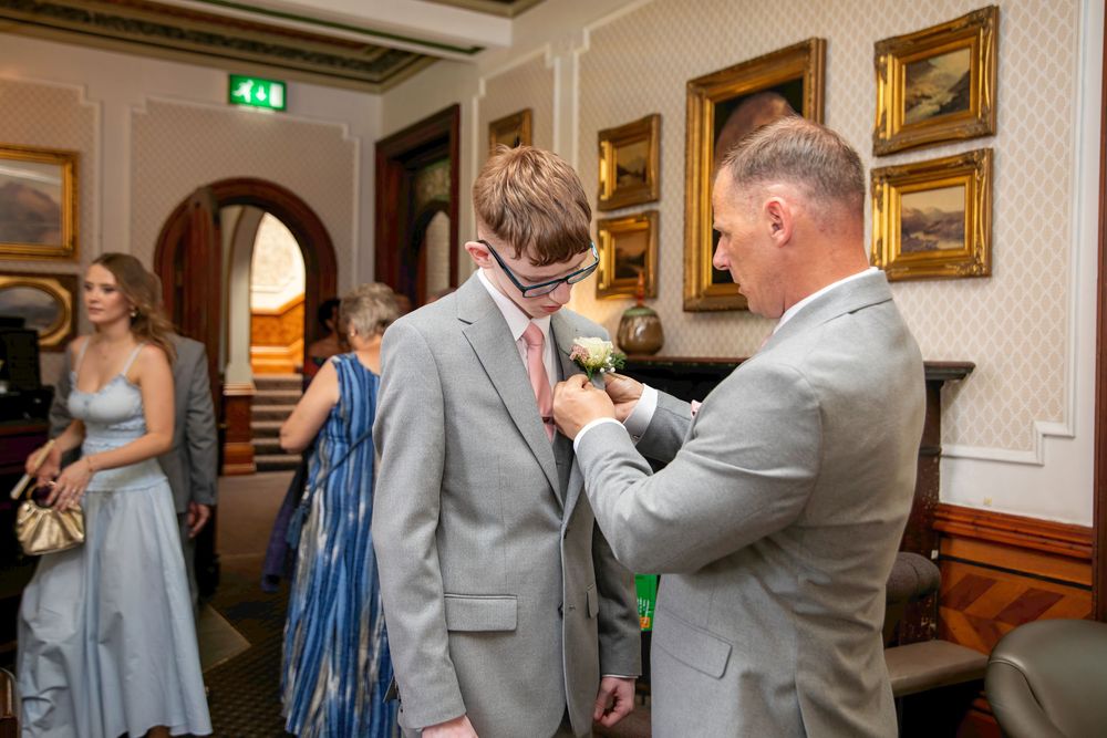 A man in a gray suit having a boutonniere pinned to his lapel at a formal wedding venue.