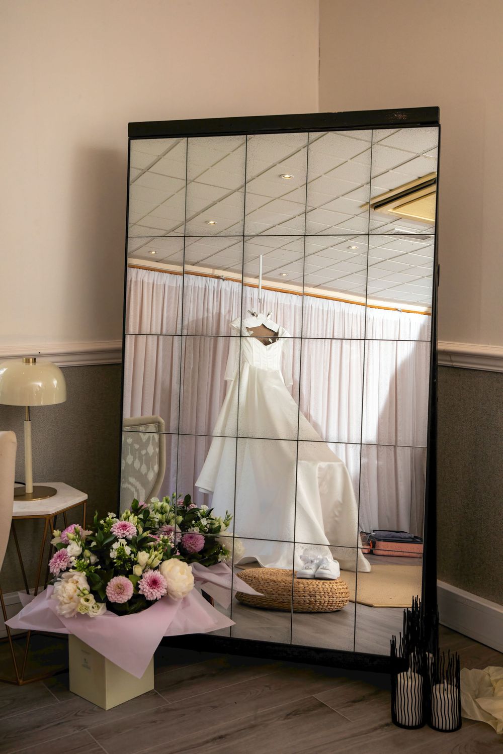 A large tiled mirror reflects a white wedding dress hanging in a bright room, with a pink floral bouquet in the foreground.