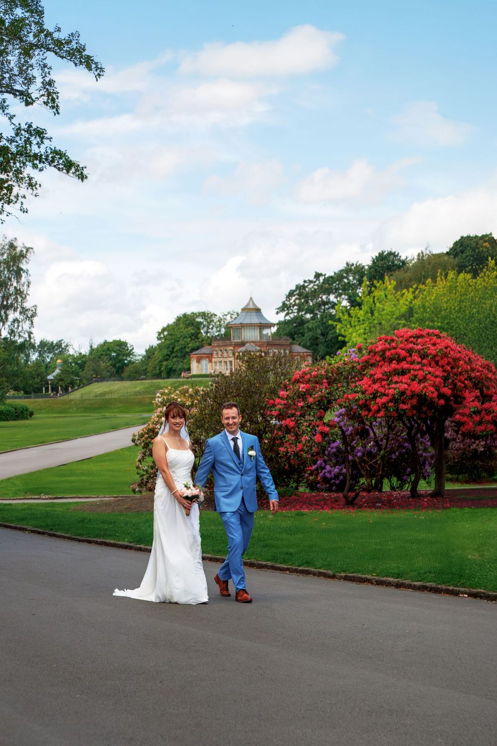 Bride and groom walking hand in hand along a path with colorful gardens and a mansion behind them.