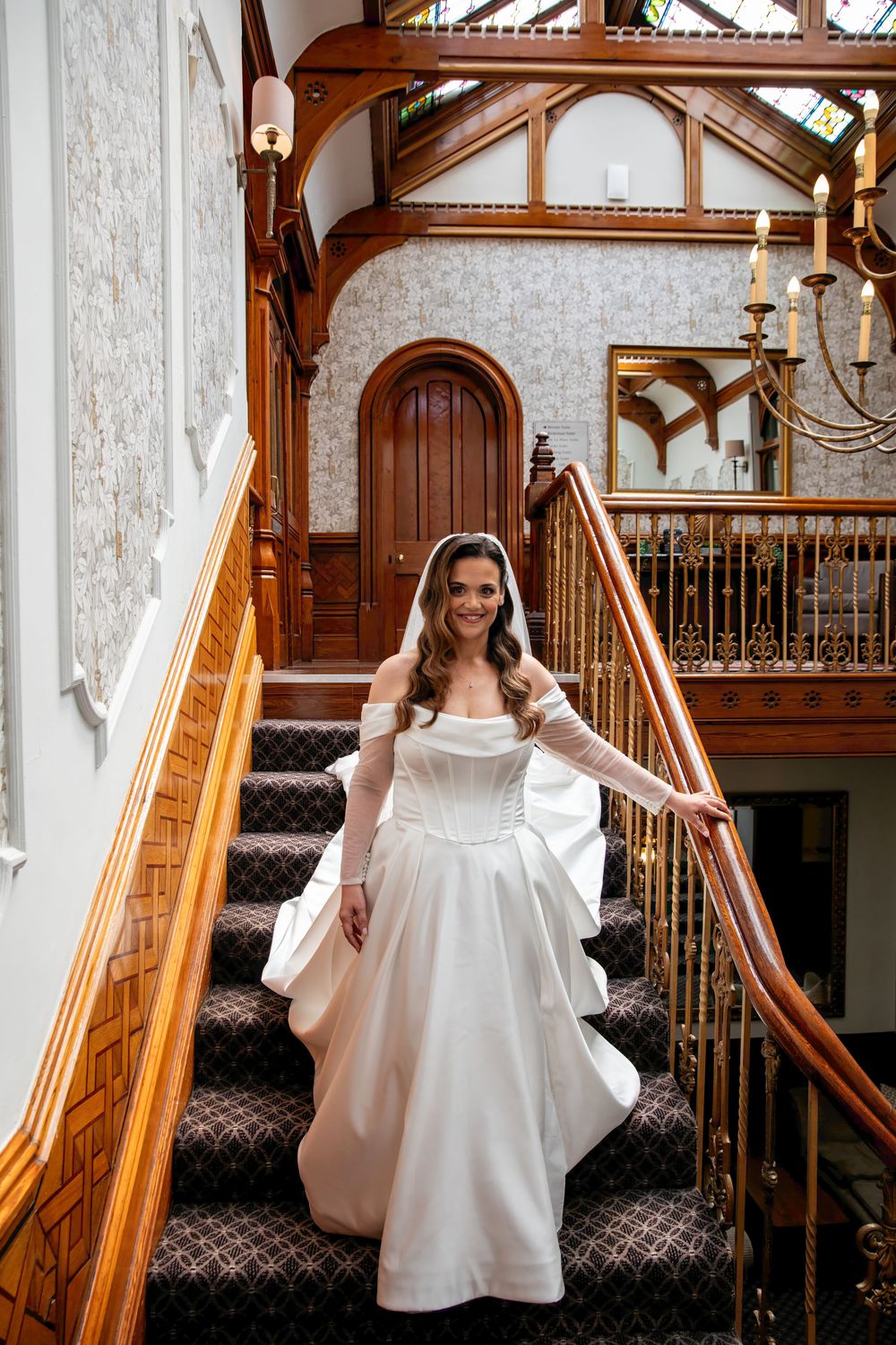 Bride in white gown and veil descends ornate wooden staircase in elegant Victorian mansion.