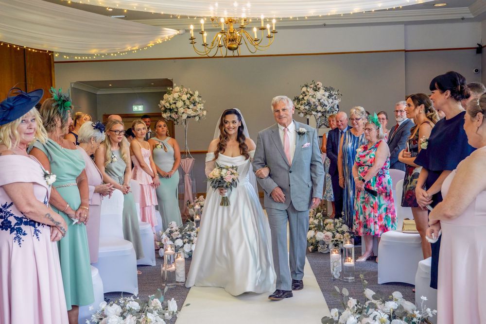 A bride walks down the aisle with her father during an elegant wedding ceremony surrounded by guests and floral decor.