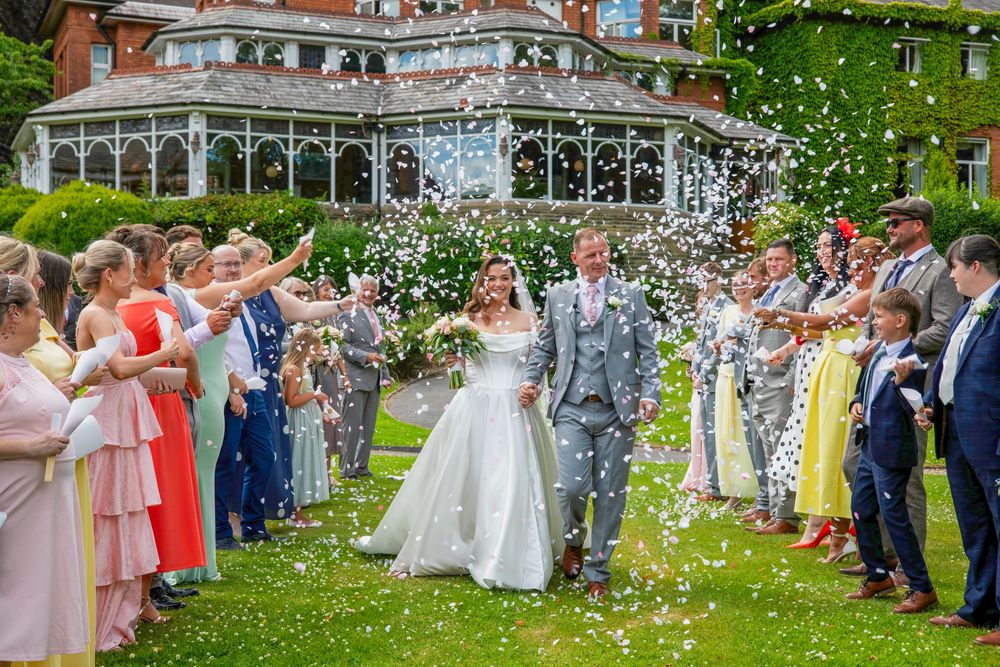 A bride and groom walk through a confetti shower surrounded by guests outside an elegant ivy-covered wedding venue.