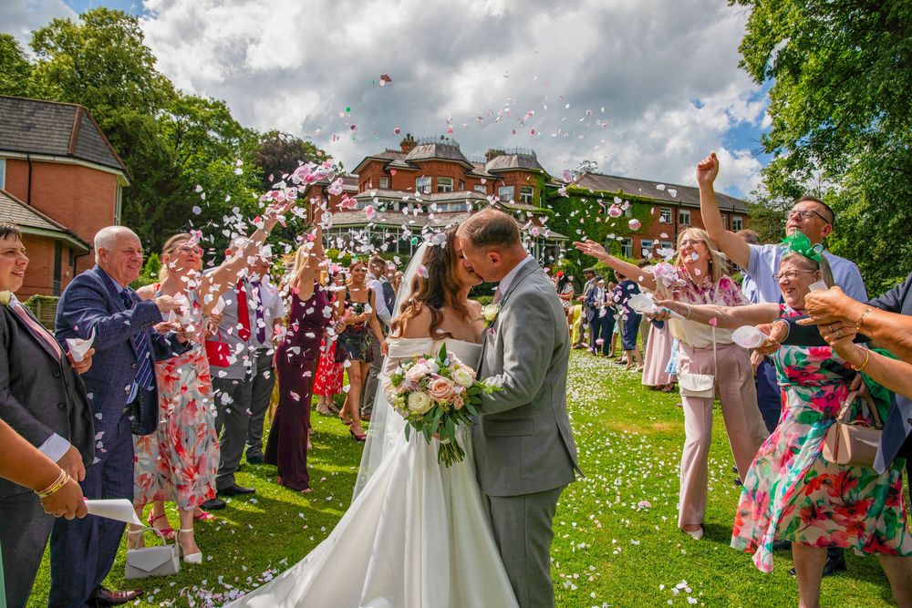 A bride and groom share a kiss as wedding guests throw confetti outdoors on a sunny day.