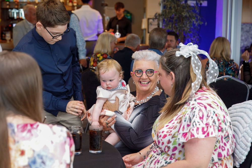 Group of people gathered at a social event, smiling woman holding a baby surrounded by family members at a table.