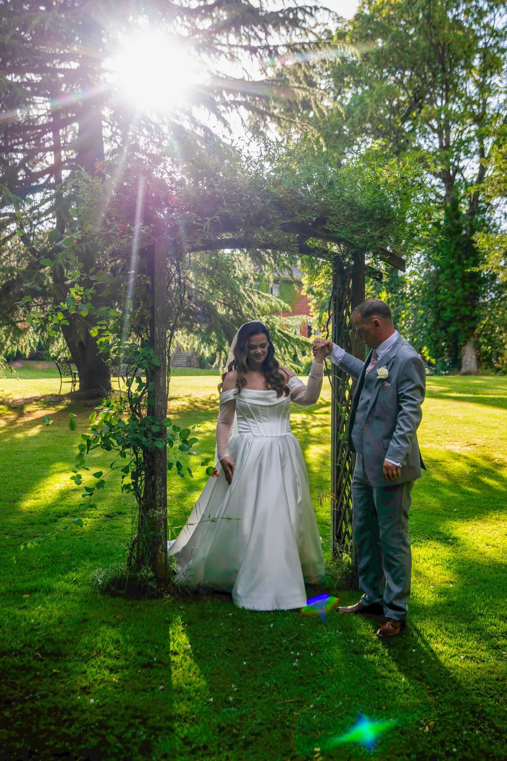 Bride and groom dancing under a garden arch in sunlight, surrounded by lush green trees on a sunny wedding day.
