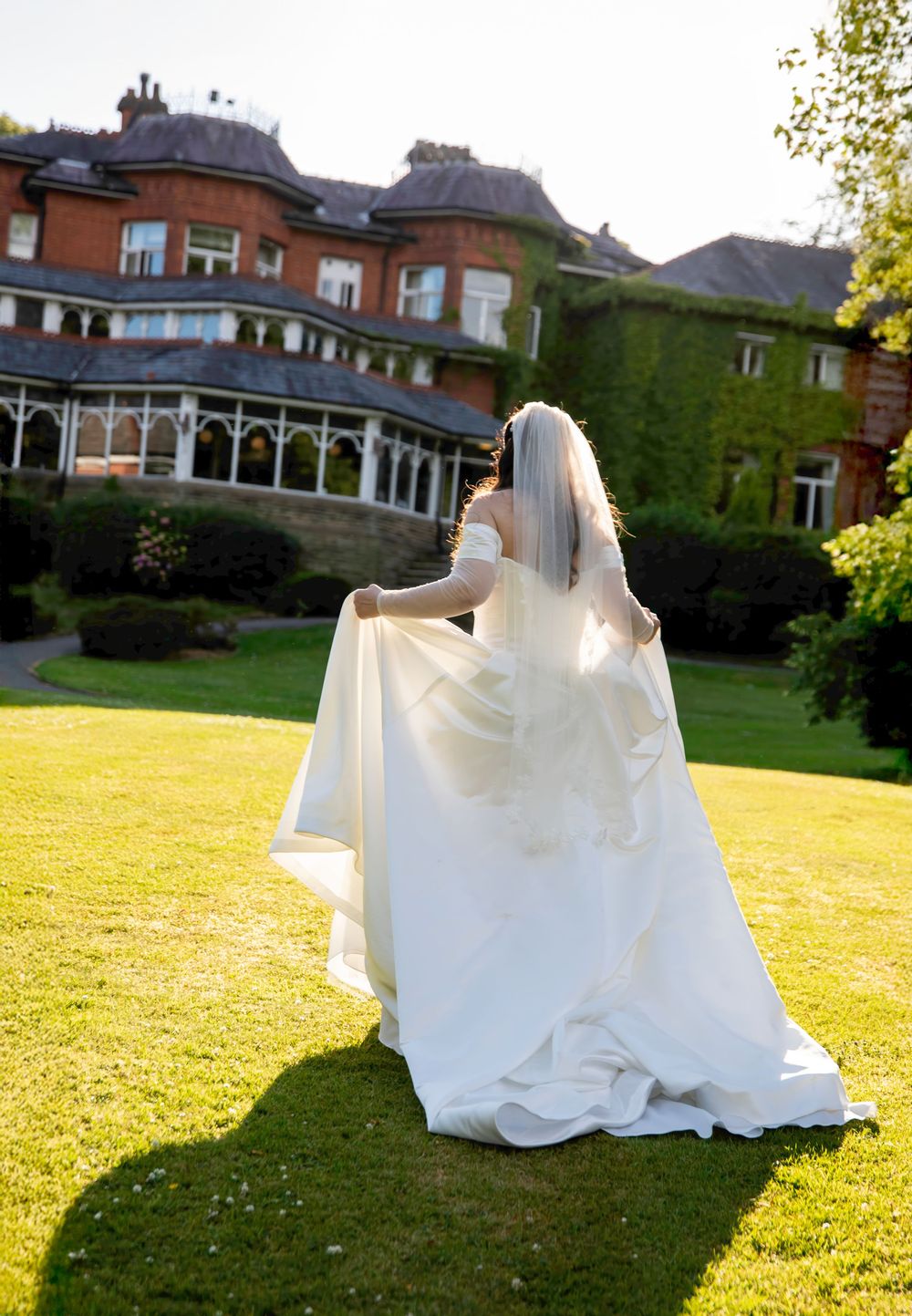 Bride in white gown and veil stands on lush green lawn before elegant red brick manor house on sunny day.