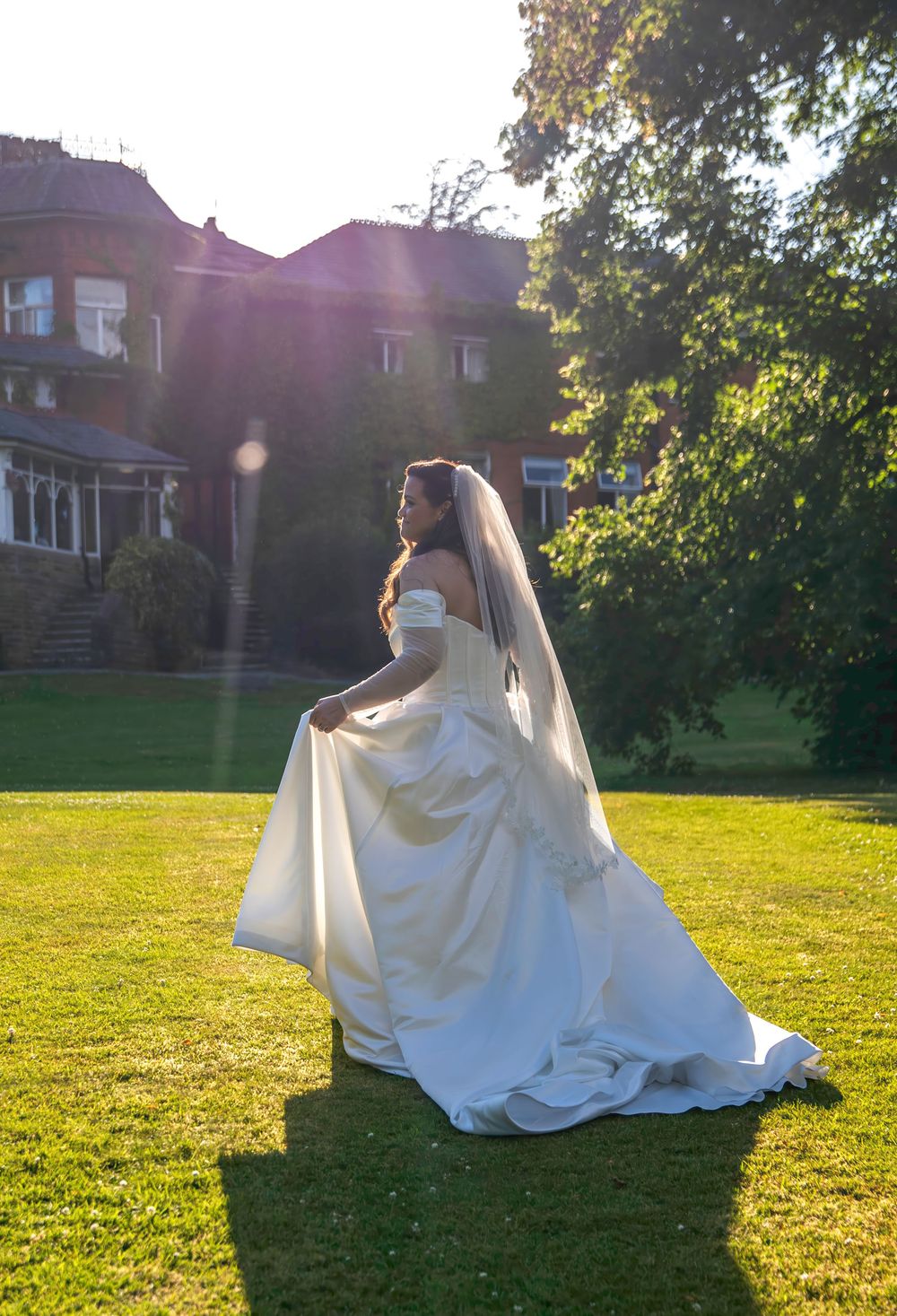 Bride in white off-shoulder gown with veil stands on green lawn before a brick manor in golden sunlight.