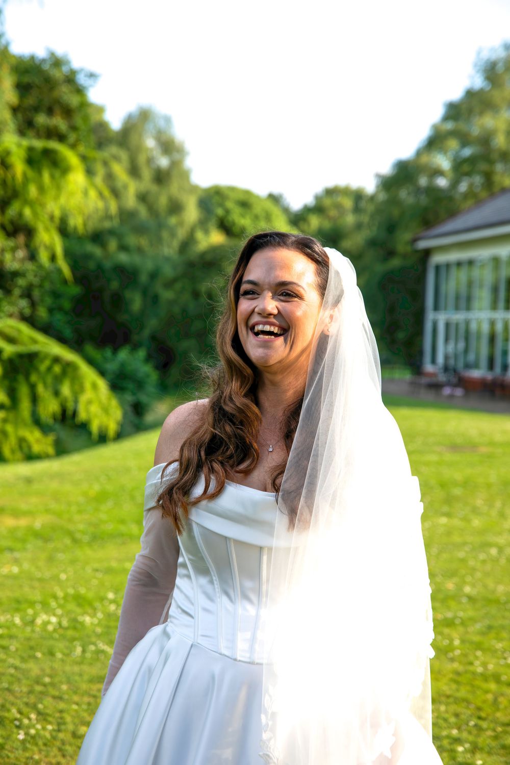 A laughing bride in a white strapless gown and veil stands on a sunny green lawn with trees and a building behind her.