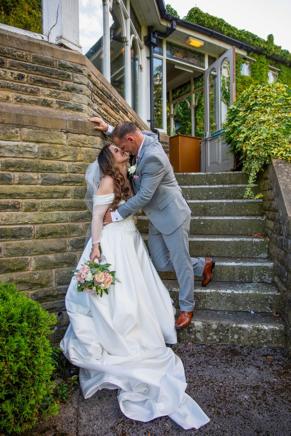 Bride and groom sharing romantic dip kiss on stone steps outside ivy-covered building, bride holding pink bouquet.