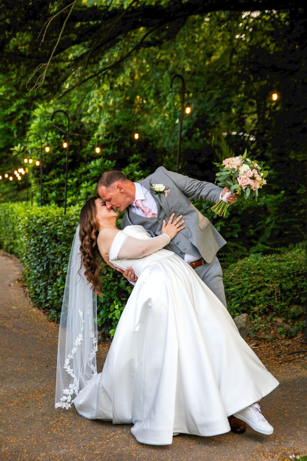 A groom dips his bride for a romantic kiss in a lush garden, her white gown and veil flowing as she holds a pink bouquet.