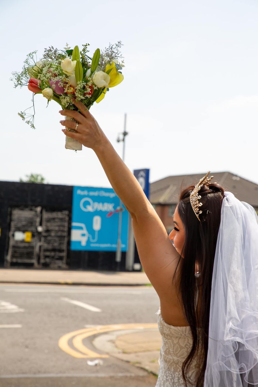 Bride in white dress holding bouquet up against blue sky.