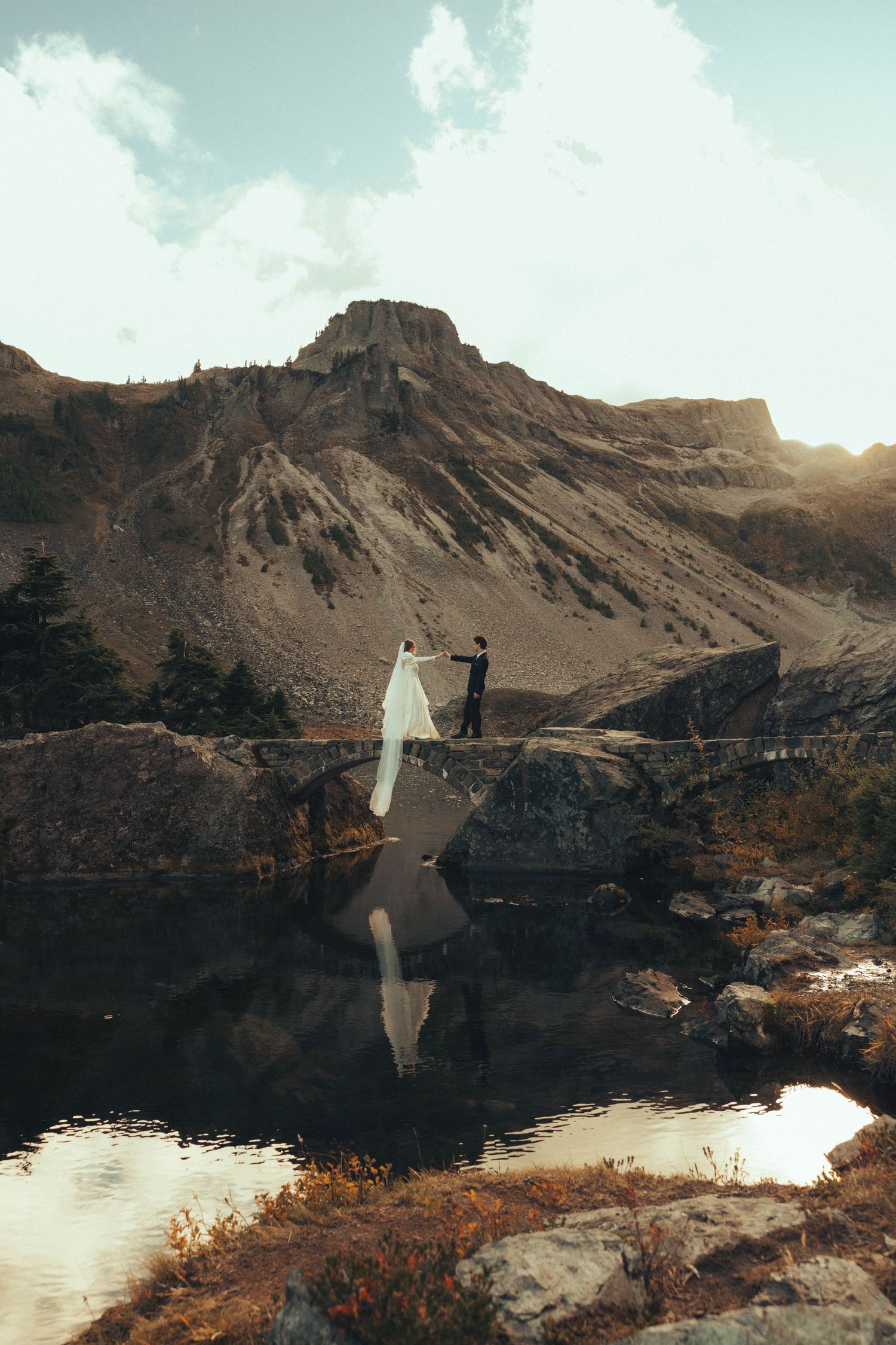 Bride and groom holding hands on a stone bridge over a reflective mountain lake during a scenic outdoor wedding.
