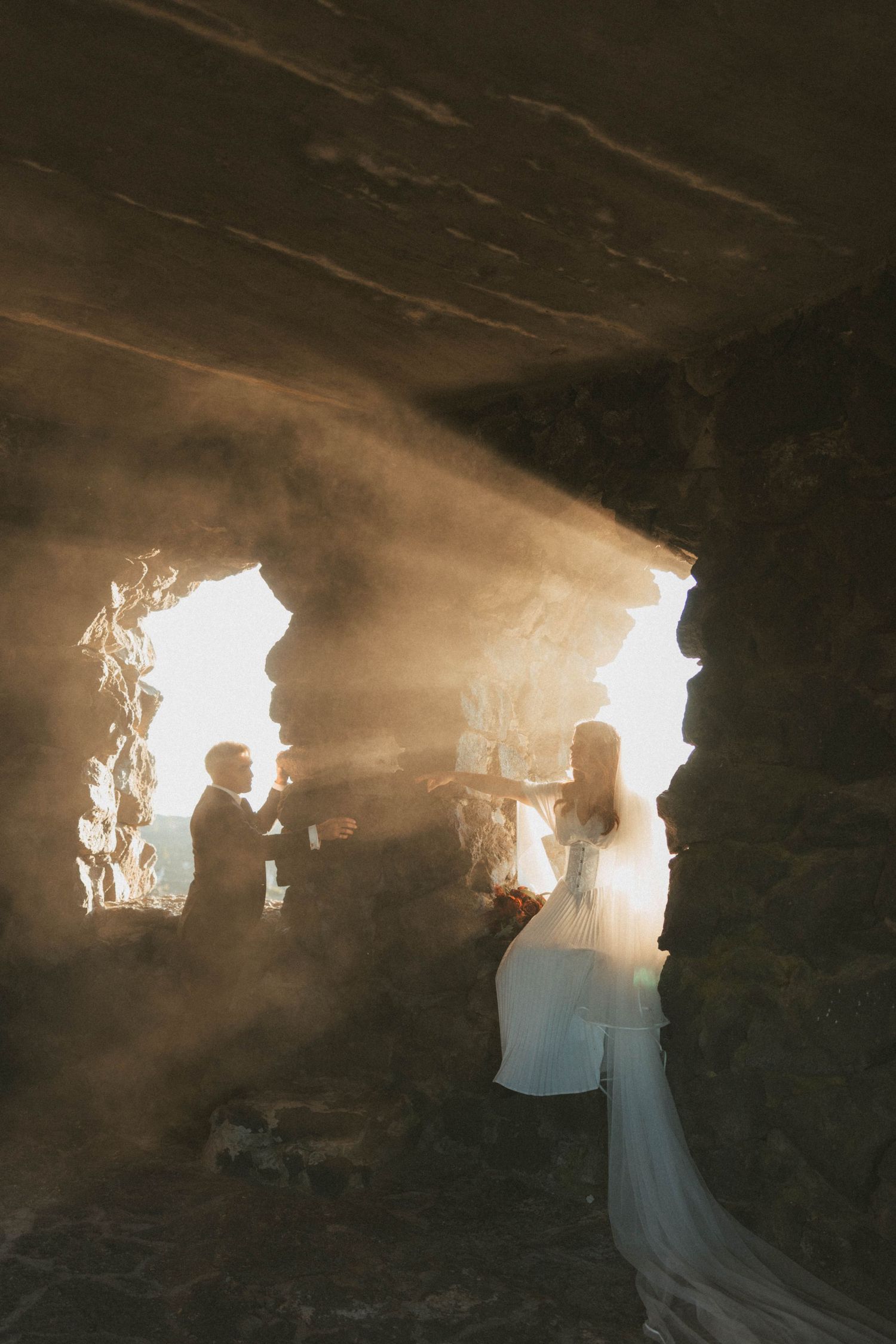 Bride and groom exchange vows in a dramatic stone ruin with golden sunbeams streaming through ancient windows.