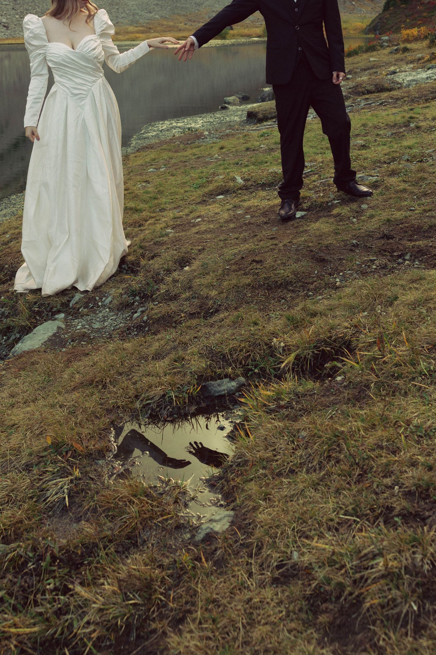 Bride in white gown and groom in black suit hold hands outdoors, reflected in a puddle on mossy ground.
