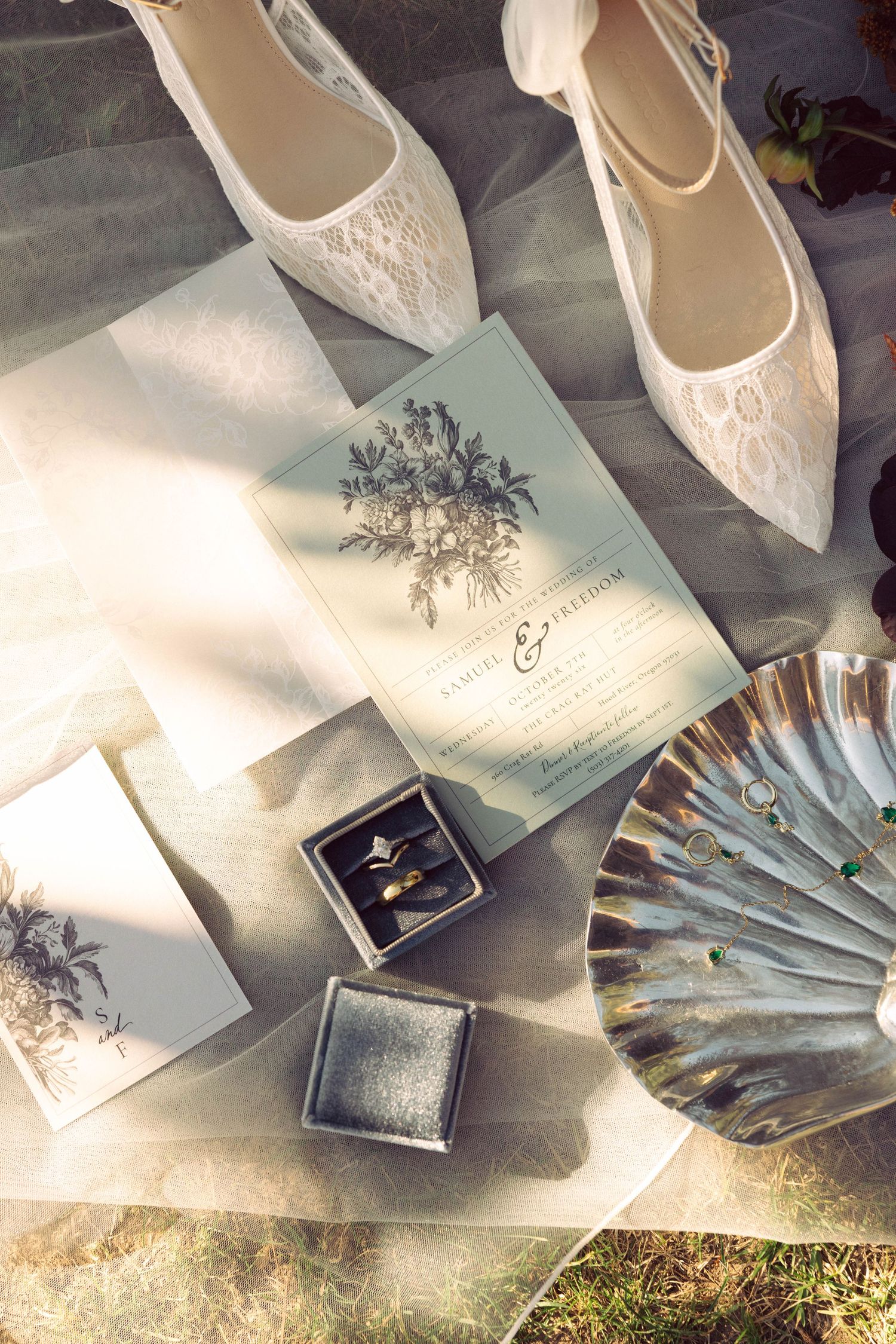 Elegant wedding flat lay with lace bridal shoes, floral invitations, ring box with gold bands, and silver shell dish.