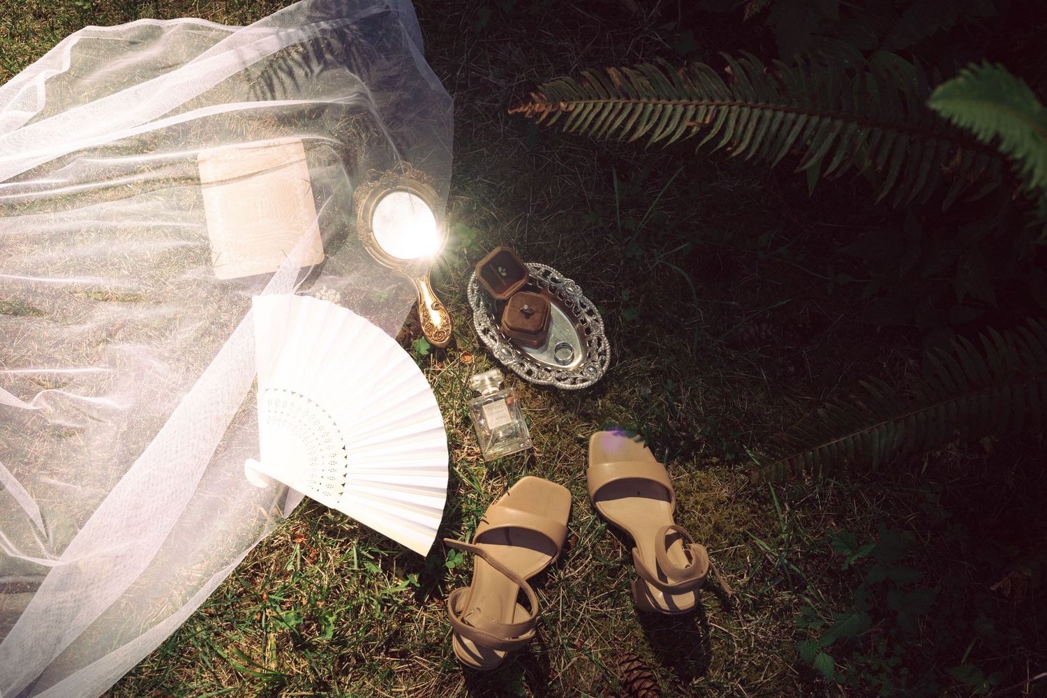 Flat lay of wedding accessories on grass at night, including a hand fan, sandals, veil, and glowing lantern.