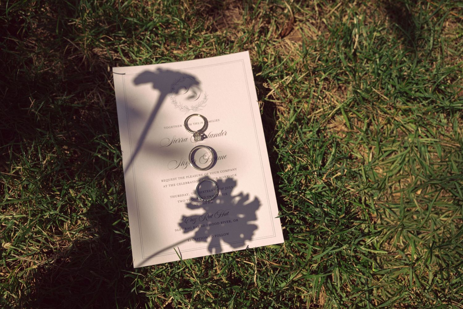 Two wedding rings resting on a white card in green grass, with flower shadows cast across the surface.