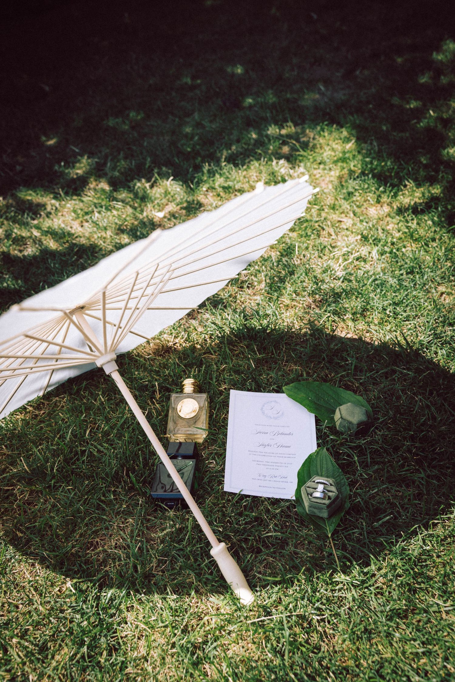 White paper parasol, perfume bottle, wedding invitation, and ring box arranged on lush green grass in soft sunlight.
