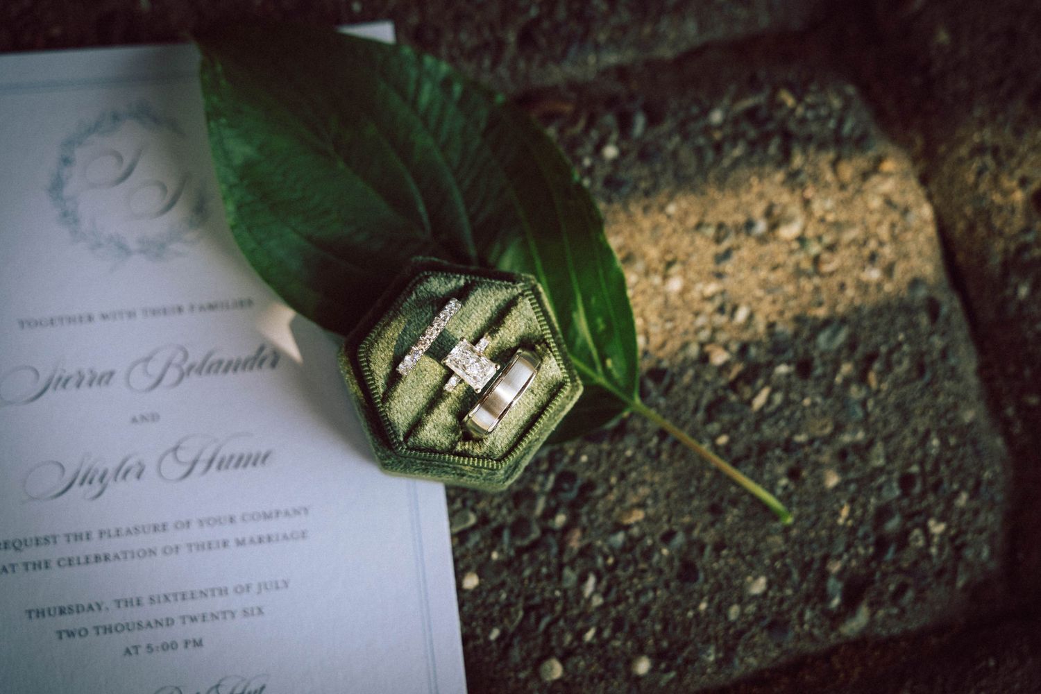 Wedding rings displayed in a green velvet hexagon box on a leaf beside a wedding invitation.