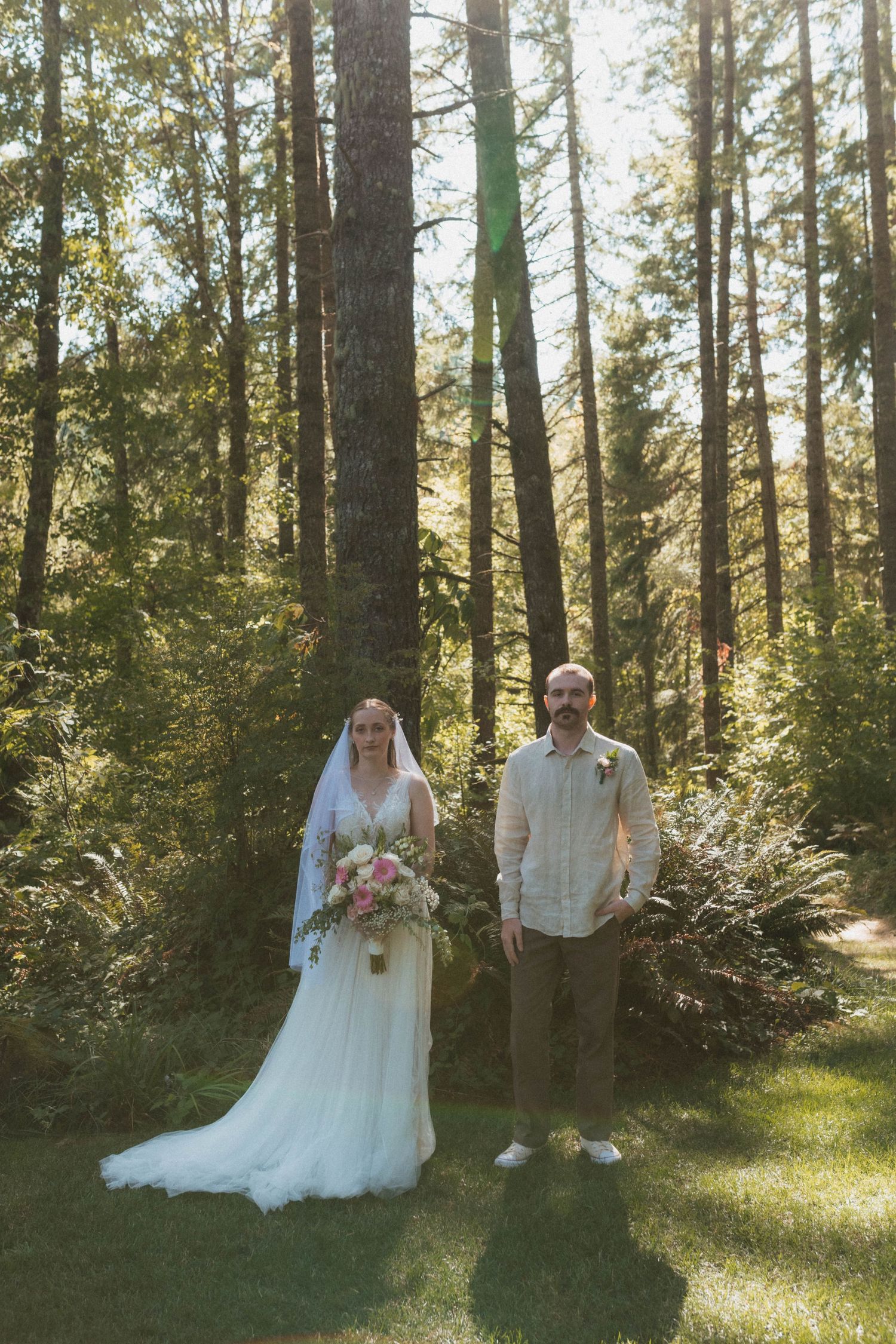 Bride in white gown with veil holding bouquet stands beside groom in tan shirt among tall trees in sunlit forest.