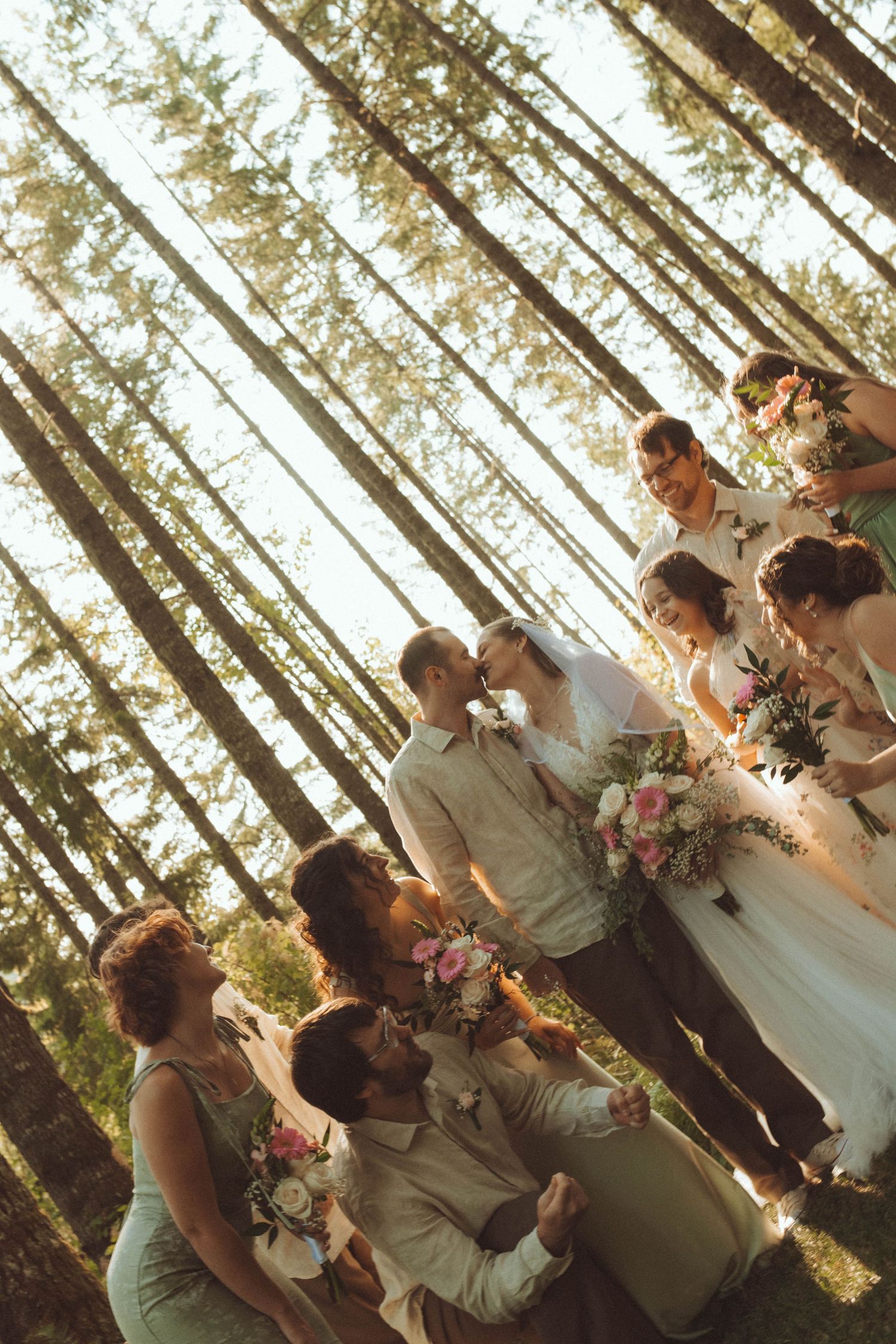 Wedding party celebrating in a sunlit forest, bride and groom kissing surrounded by bridesmaids and groomsmen holding floral bouquets.