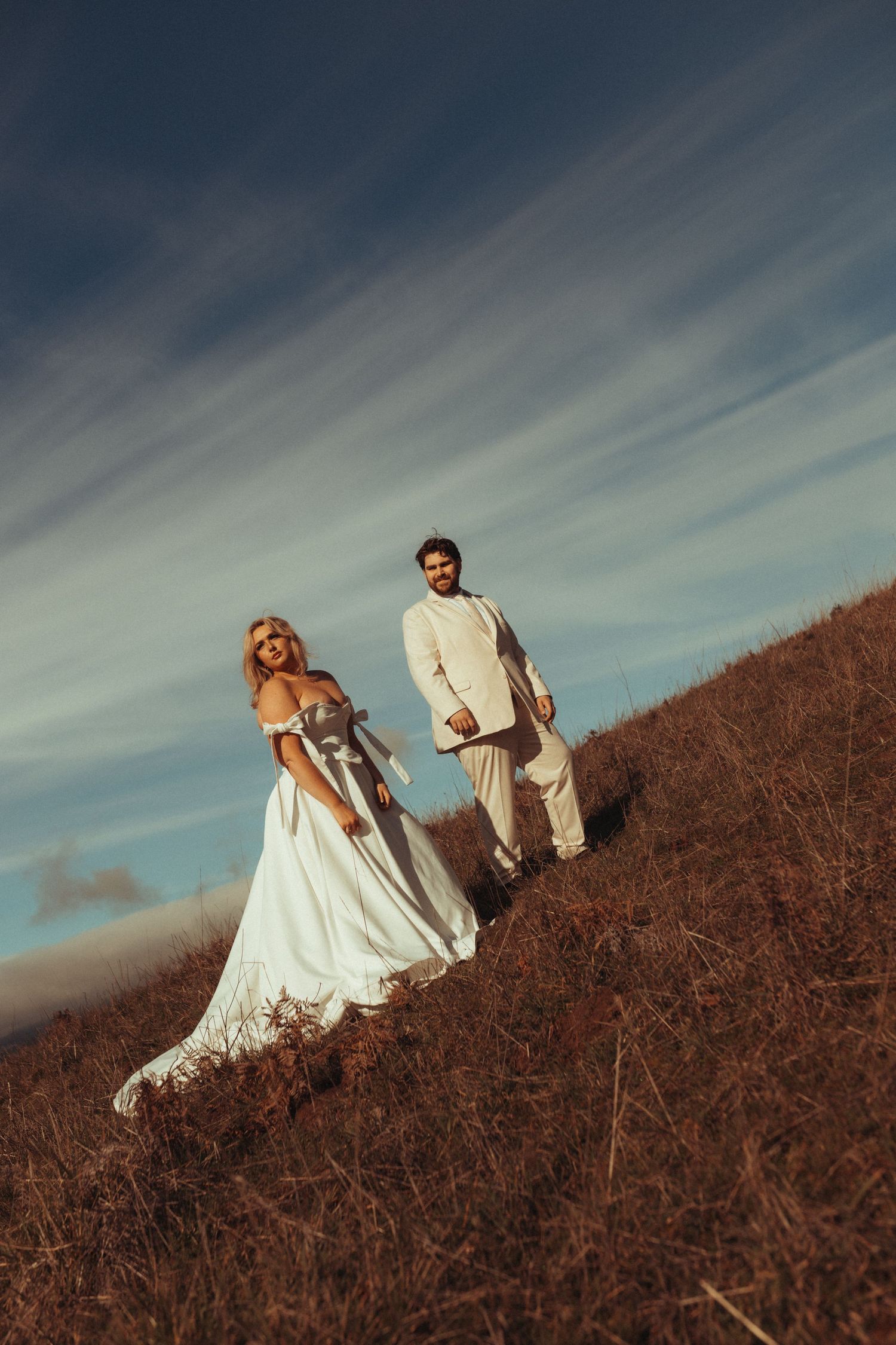 A couple in wedding attire stands on a hillside against a dramatic cloudy sky during golden hour.