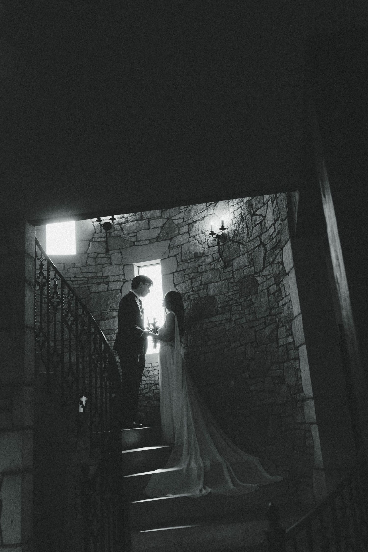 Black and white wedding photo of bride and groom silhouetted on stone staircase in romantic candlelit castle.