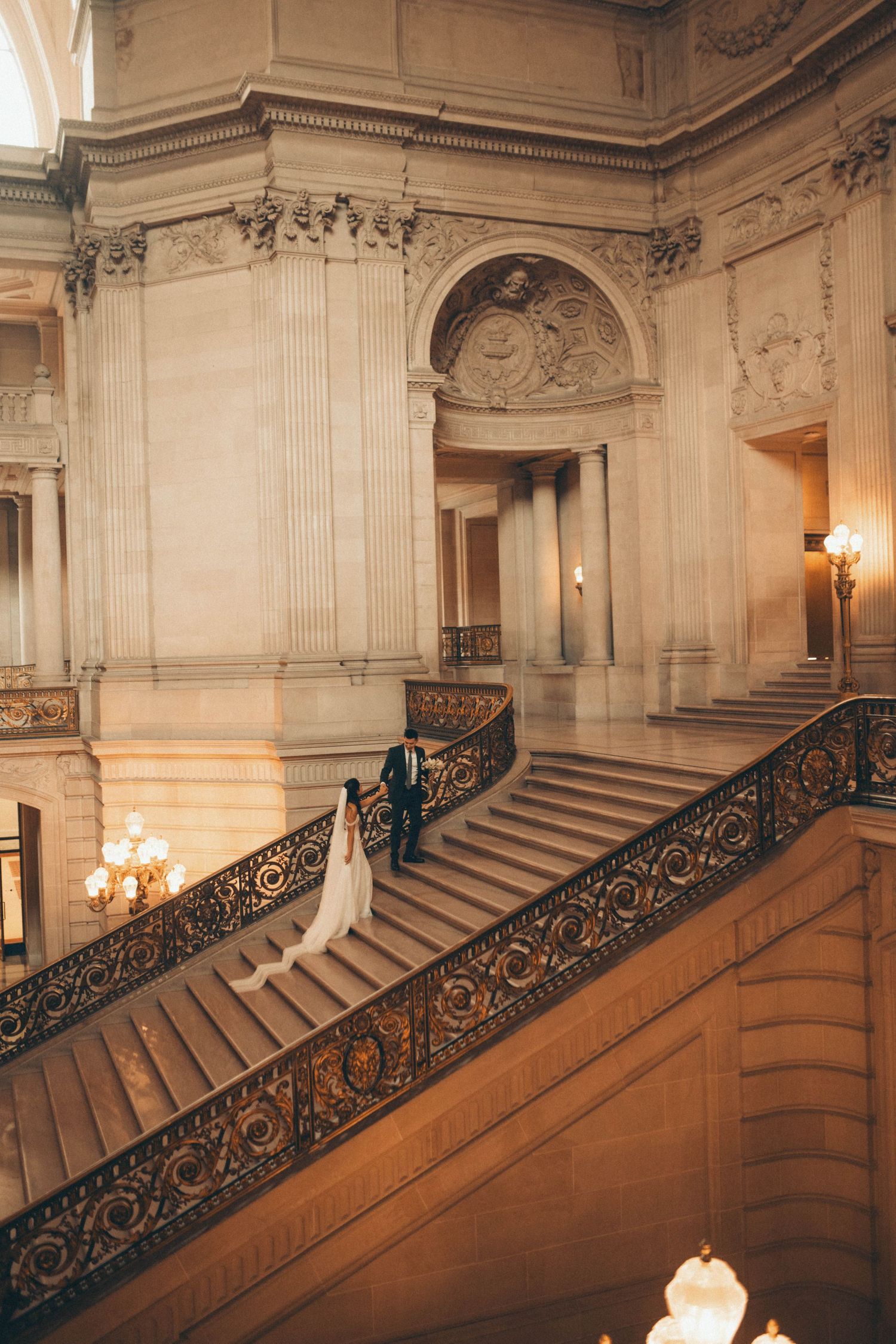 Bride and groom walking down ornate grand staircase in San Francisco City Hall during wedding ceremony.