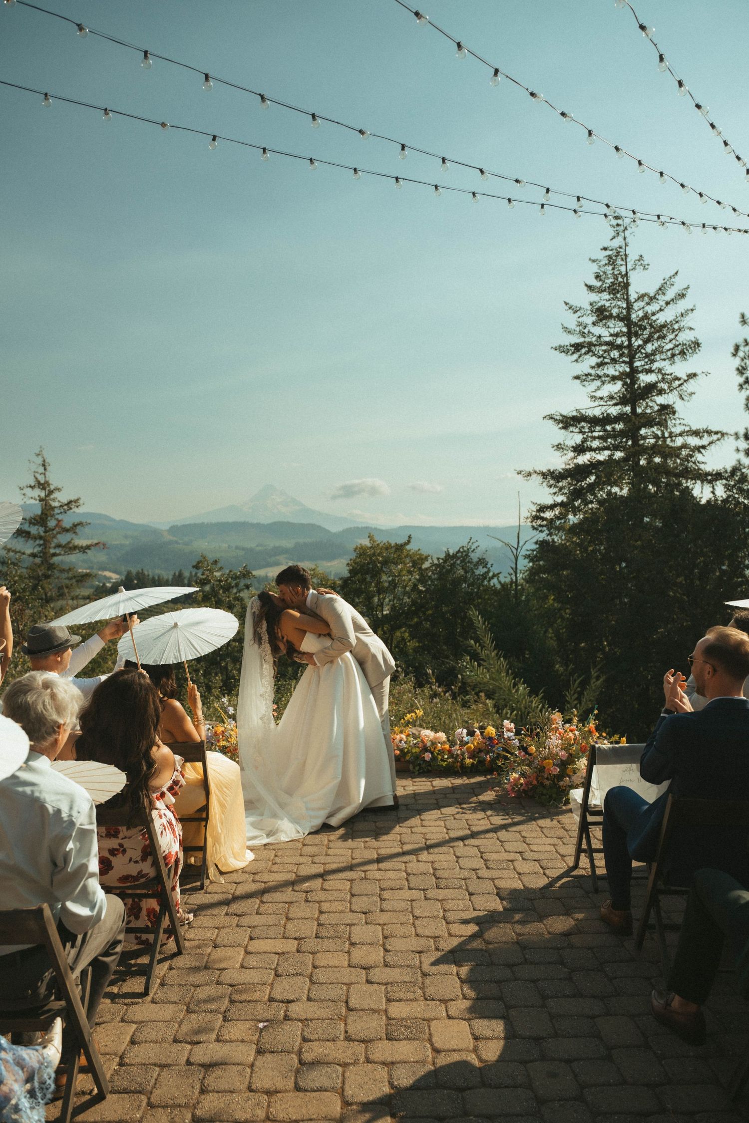 Couple sharing first kiss at outdoor wedding ceremony with mountain views, string lights, and guests holding parasols.