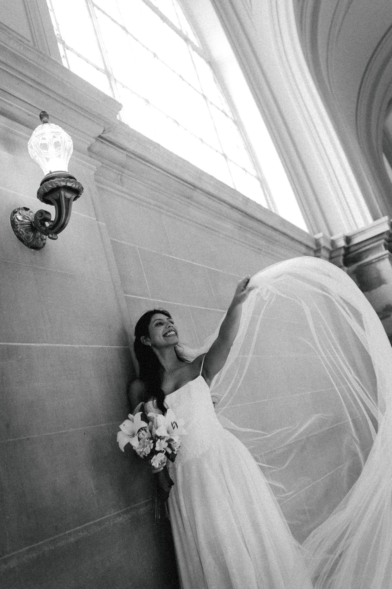 Black and white photo of a smiling bride in a flowing gown and veil holding a bouquet inside an elegant building.