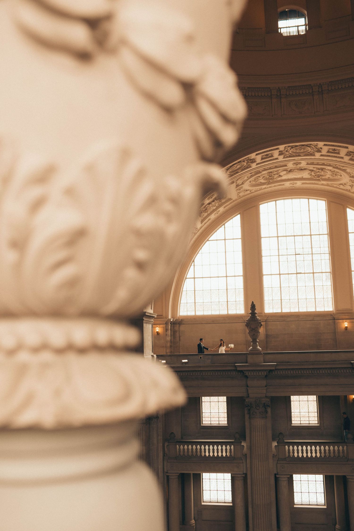 Blurred classical column foreground frames distant wedding couple on balcony of ornate domed building with arched windows.