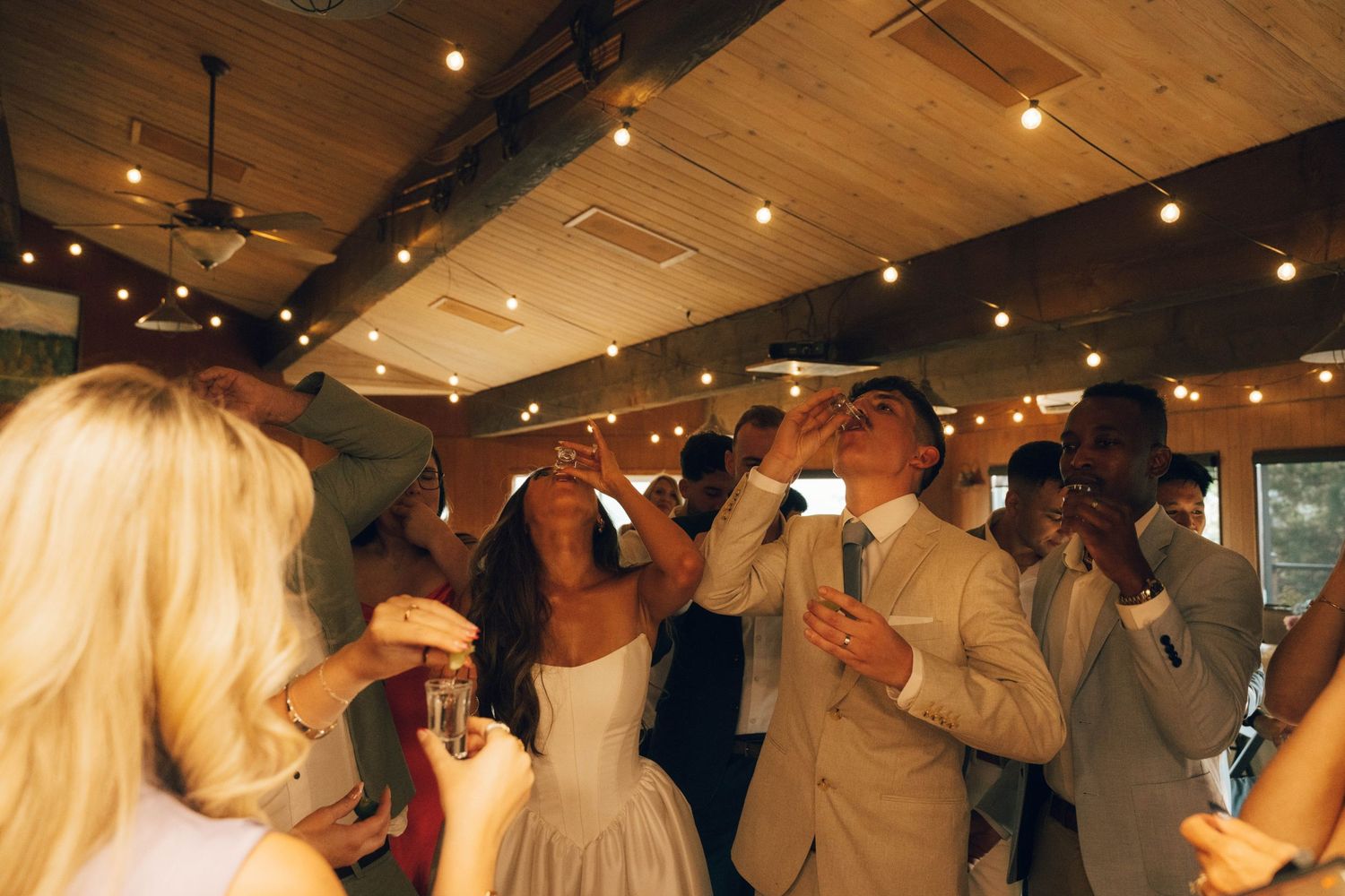 Wedding couple doing shots on dance floor surrounded by guests in elegant venue with string lights.