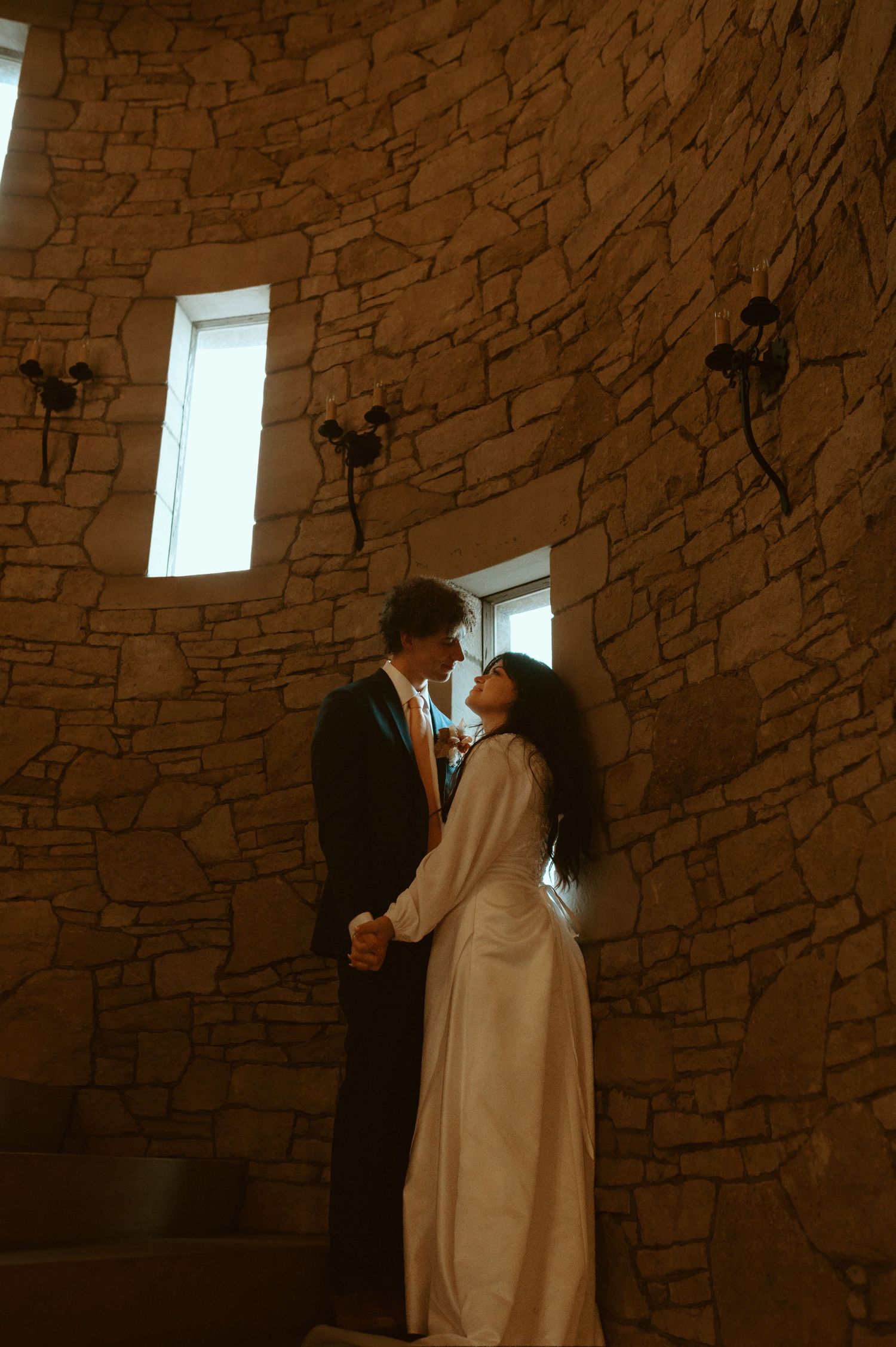 A couple stands together in an intimate moment against a rustic stone wall with high windows.