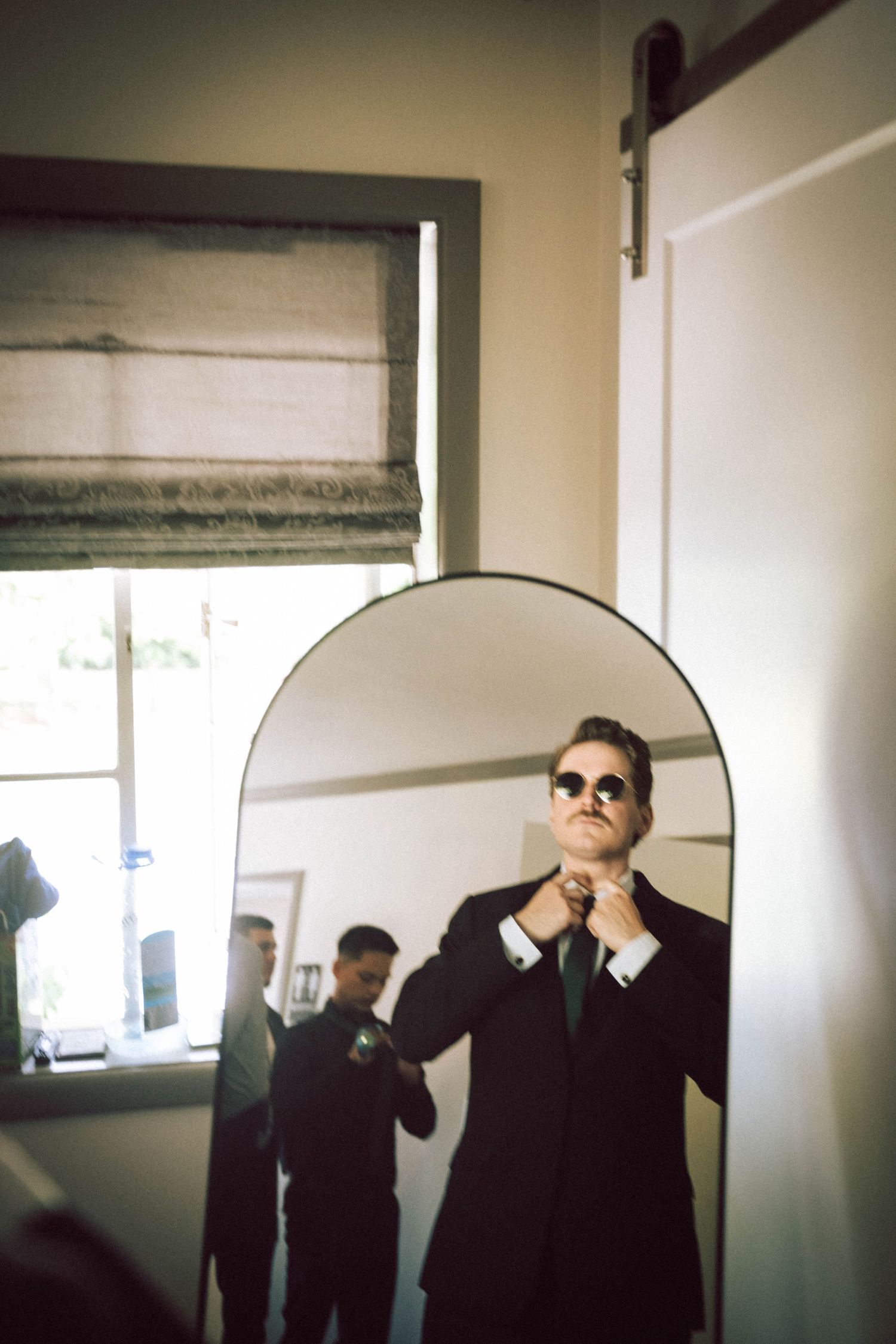 A man in a dark suit adjusts his green tie while looking in an arched mirror, with sunglasses on.
