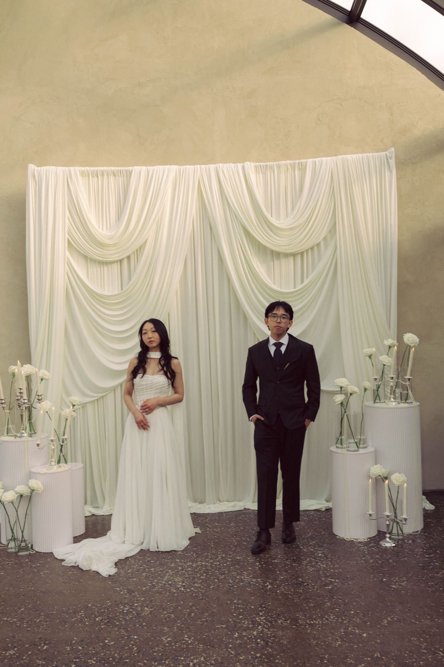 Bride in white gown and groom in black suit stand before elegant white draped backdrop with floral arrangements.