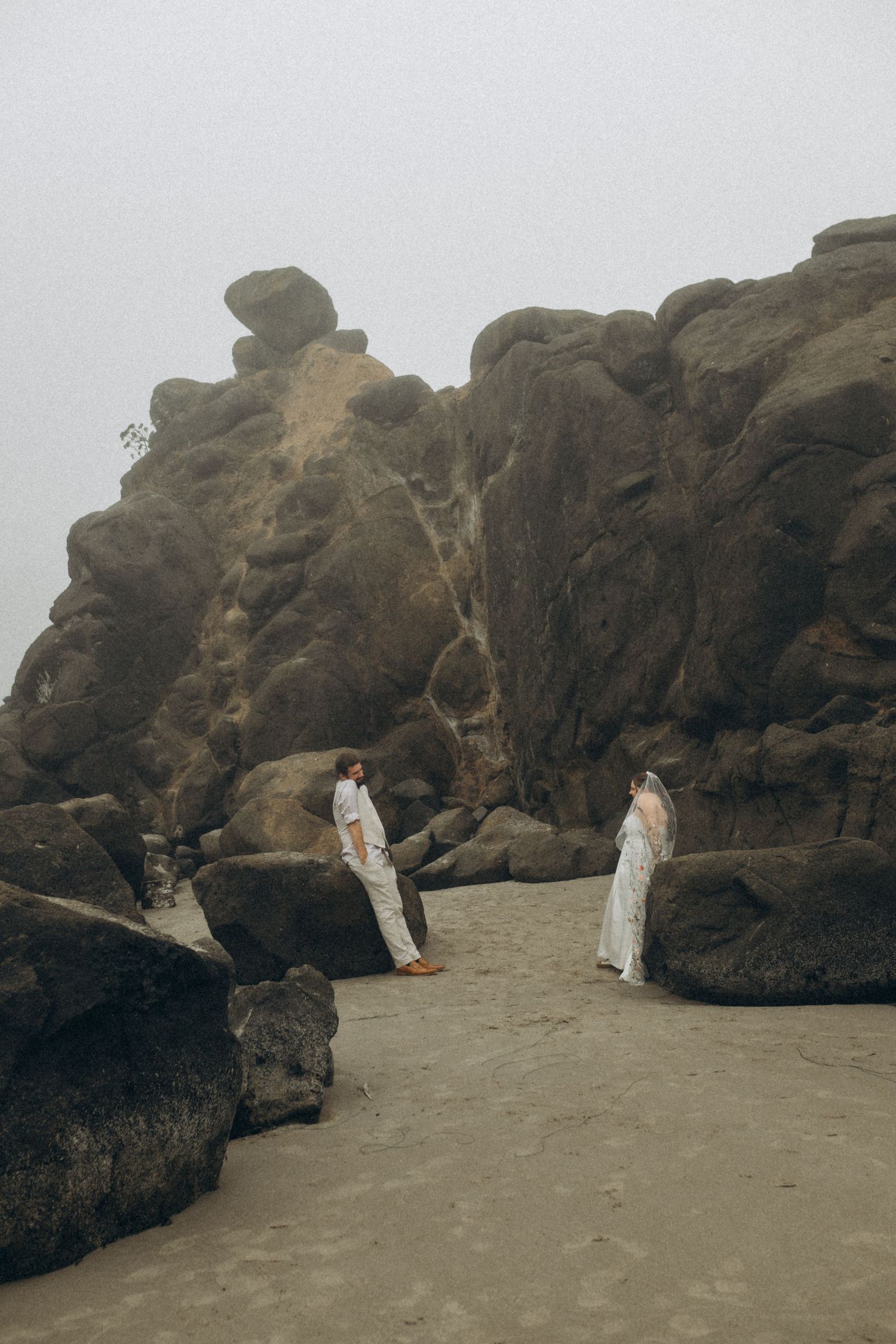 Two people in white outfits explore a rocky beach with dramatic coastal cliffs in the background.
