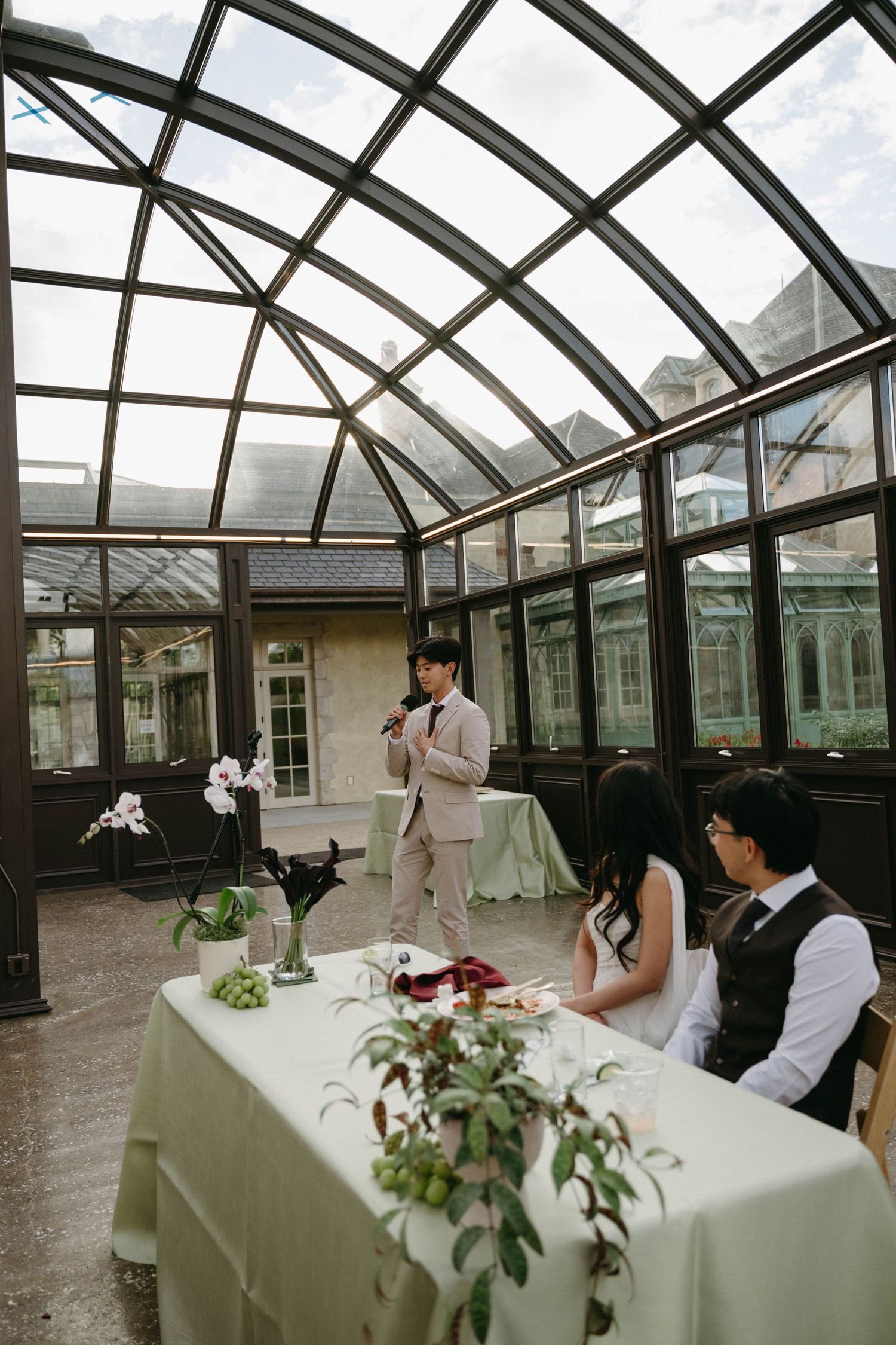A wedding reception in a glass conservatory with a man giving a speech to a seated bride and groom at a decorated table.