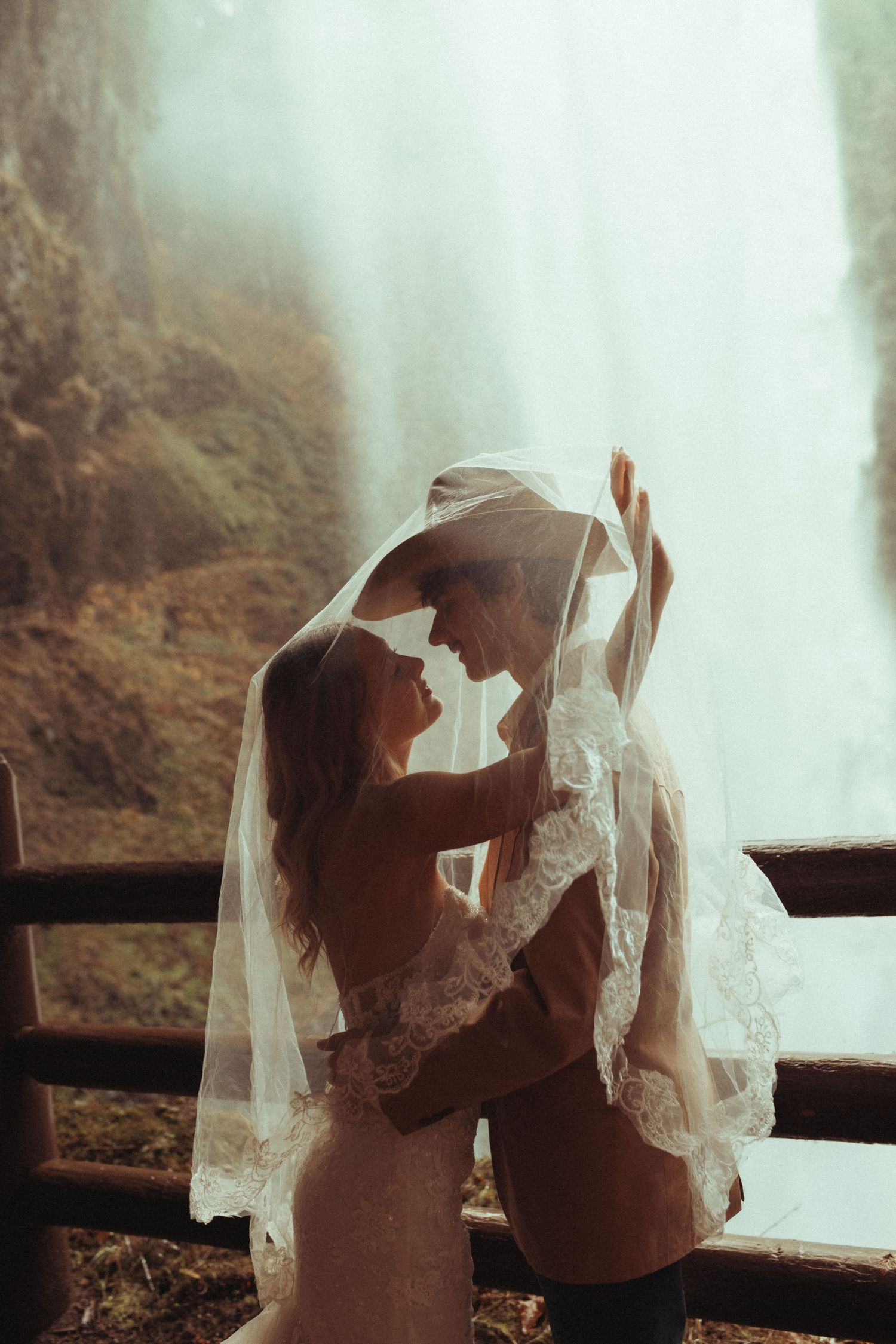 A romantic couple embraces under a delicate lace veil with a majestic waterfall cascading in the background.