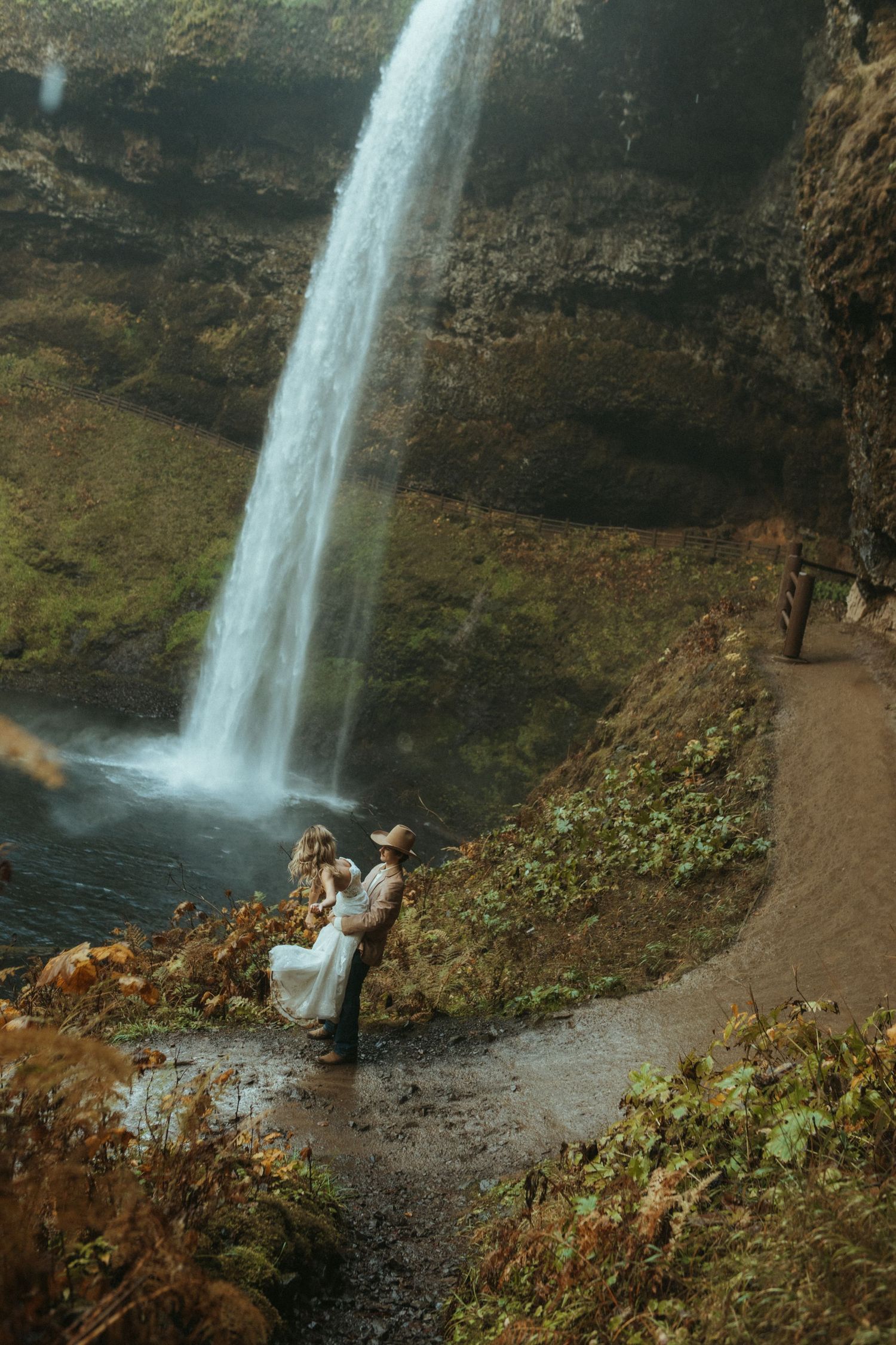 A couple shares an intimate moment on rocky terrain beside a towering waterfall in a scenic natural setting.