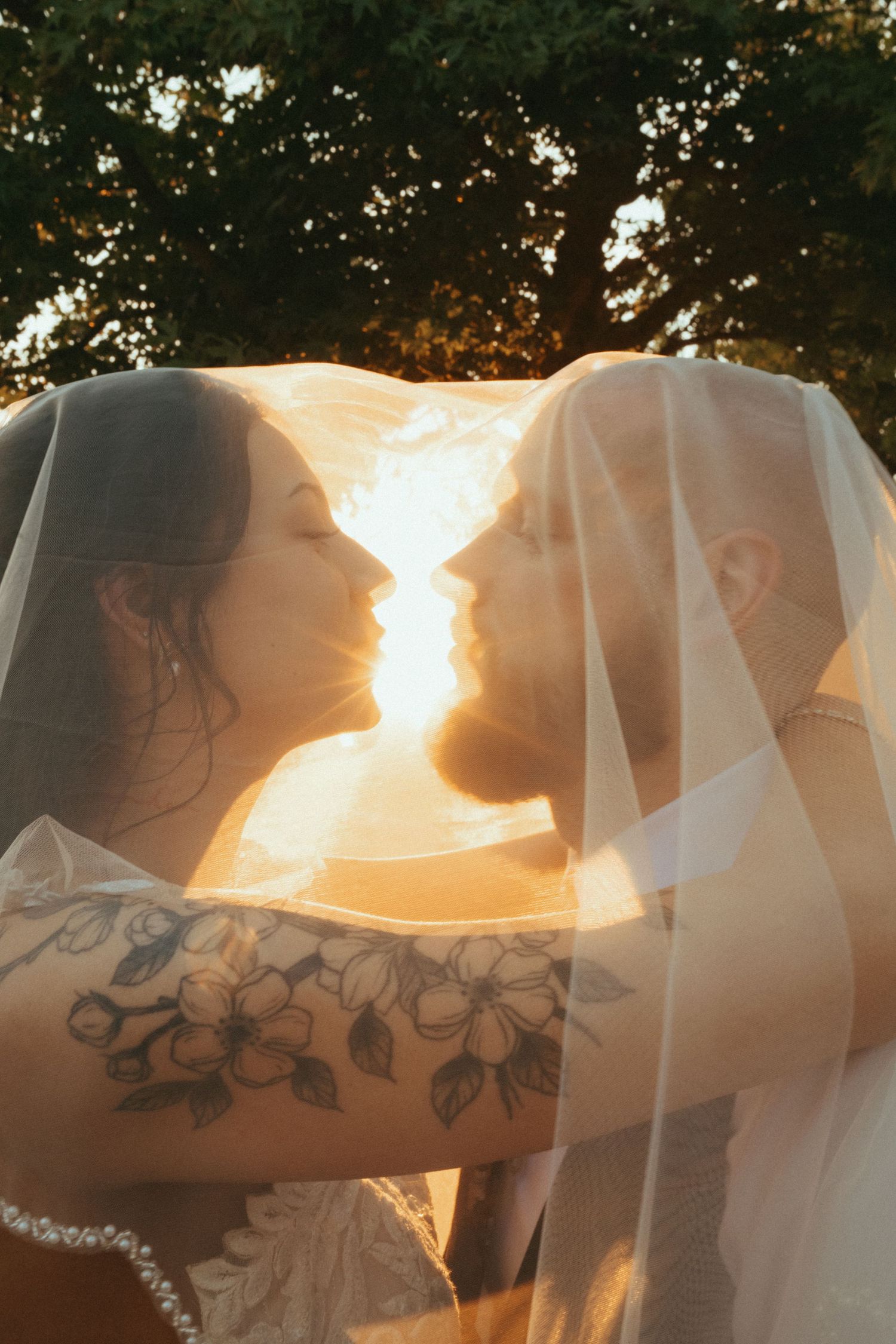 Silhouetted couple sharing a romantic kiss behind a sheer veil with sunlight glowing through at sunset.