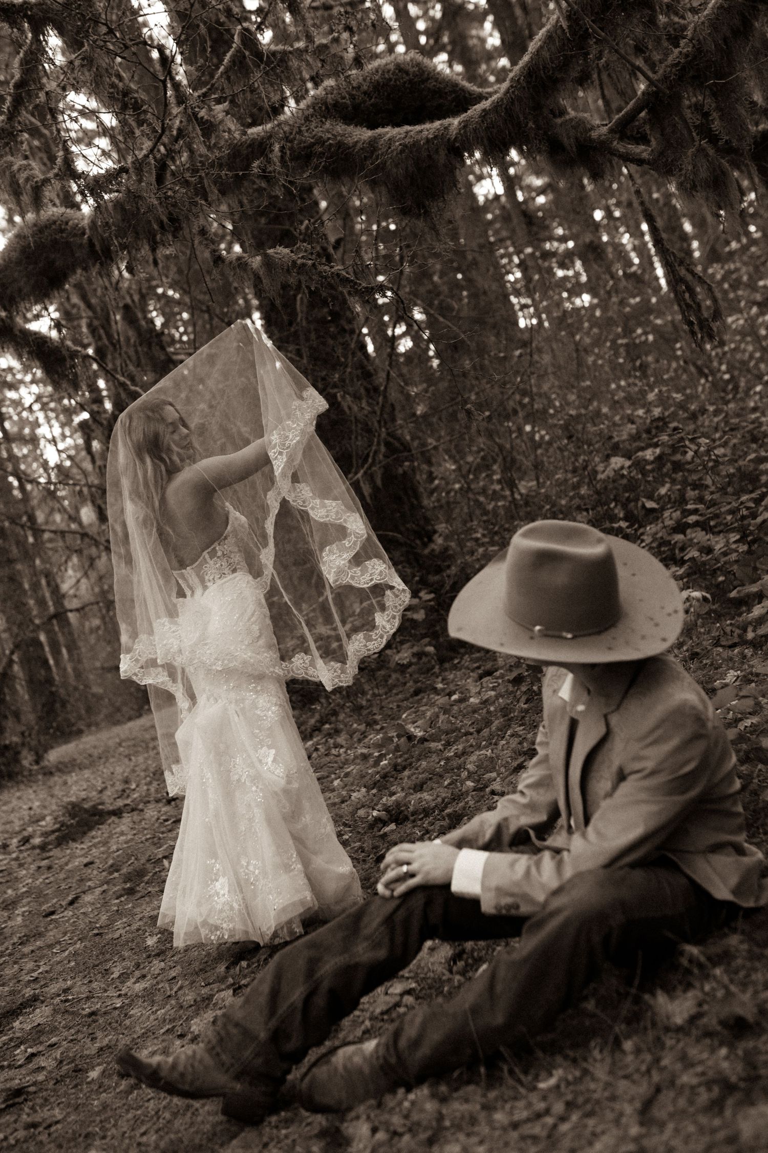 A dreamy black and white photo of a couple in vintage attire with a flowing veil in a forest setting.