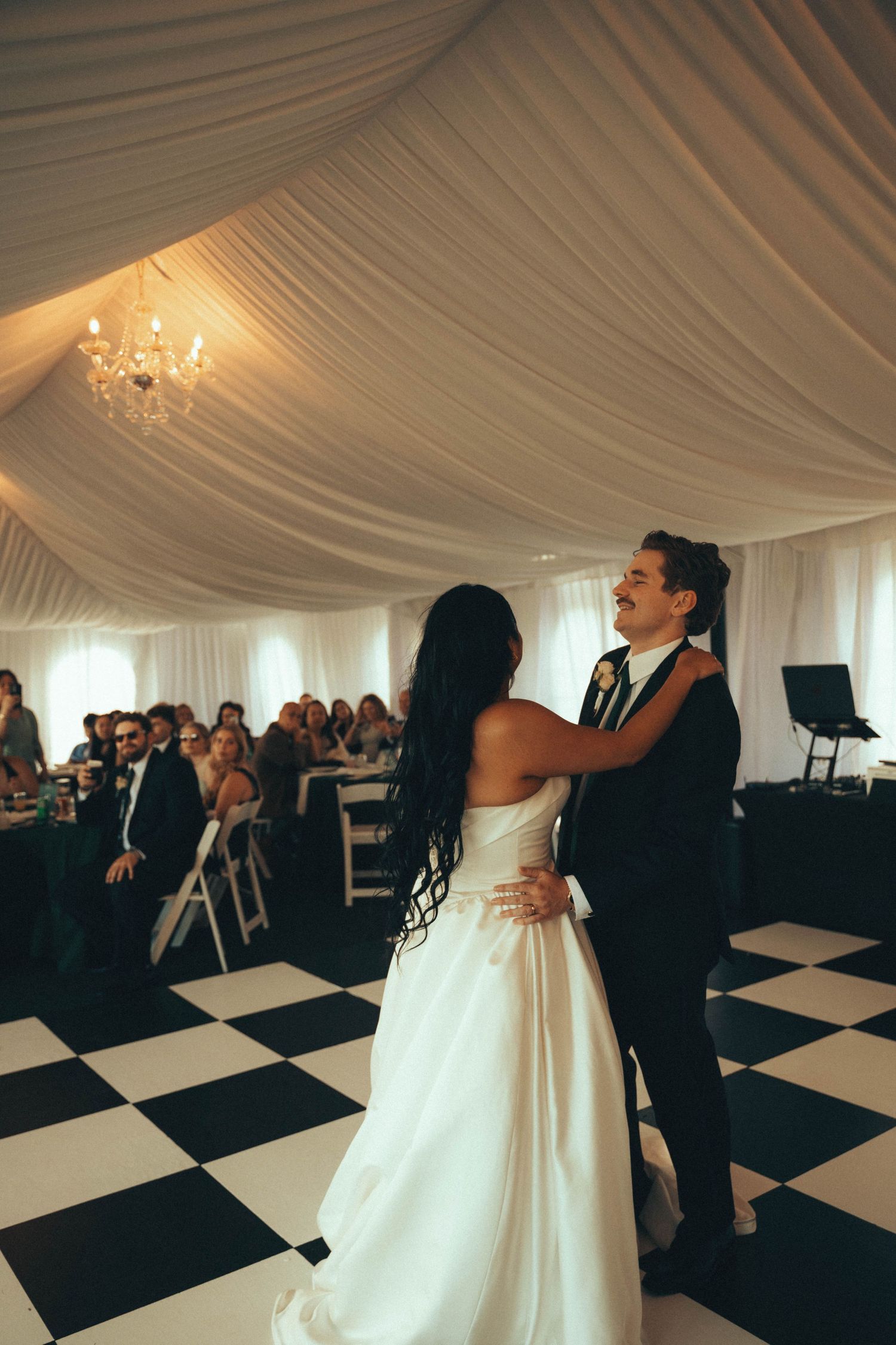 Bride and groom share their first dance on a checkered floor inside an elegant white draped wedding tent.