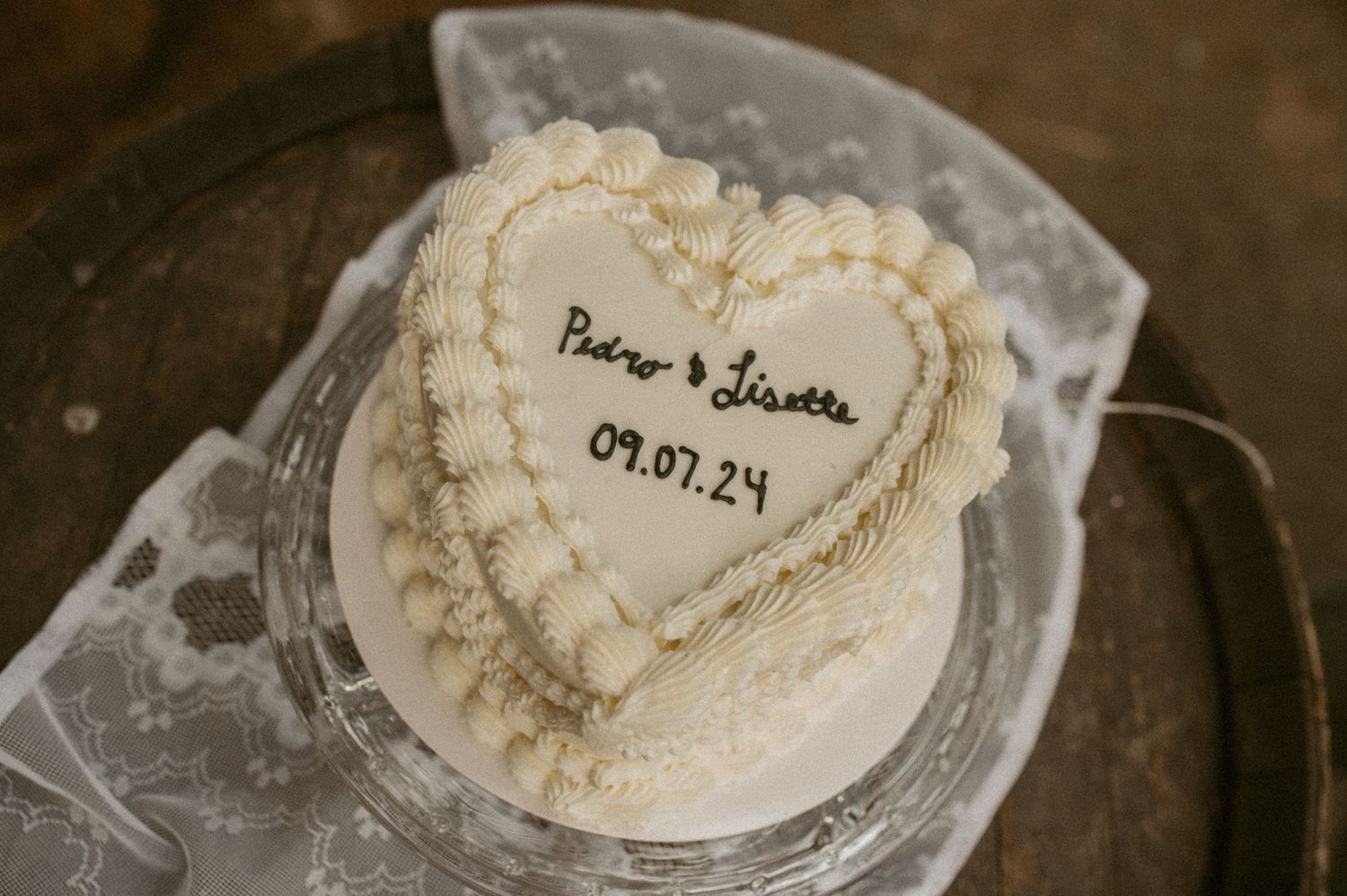 A heart-shaped white wedding cake with delicate piped border and date details sits on a glass cake stand.