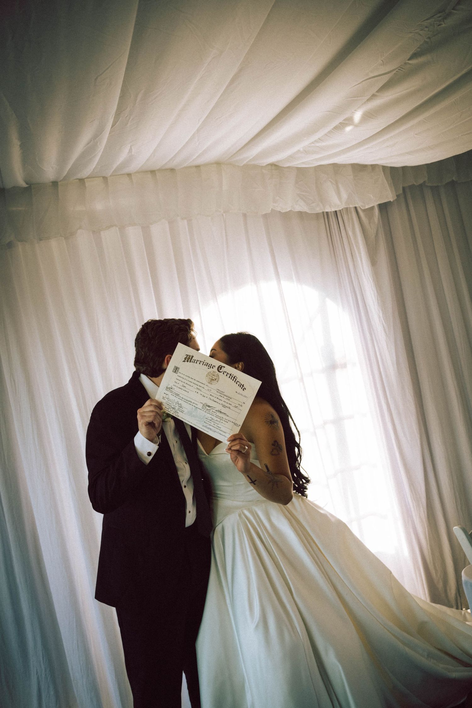 Newlyweds kissing behind their marriage certificate in a white draped wedding venue.