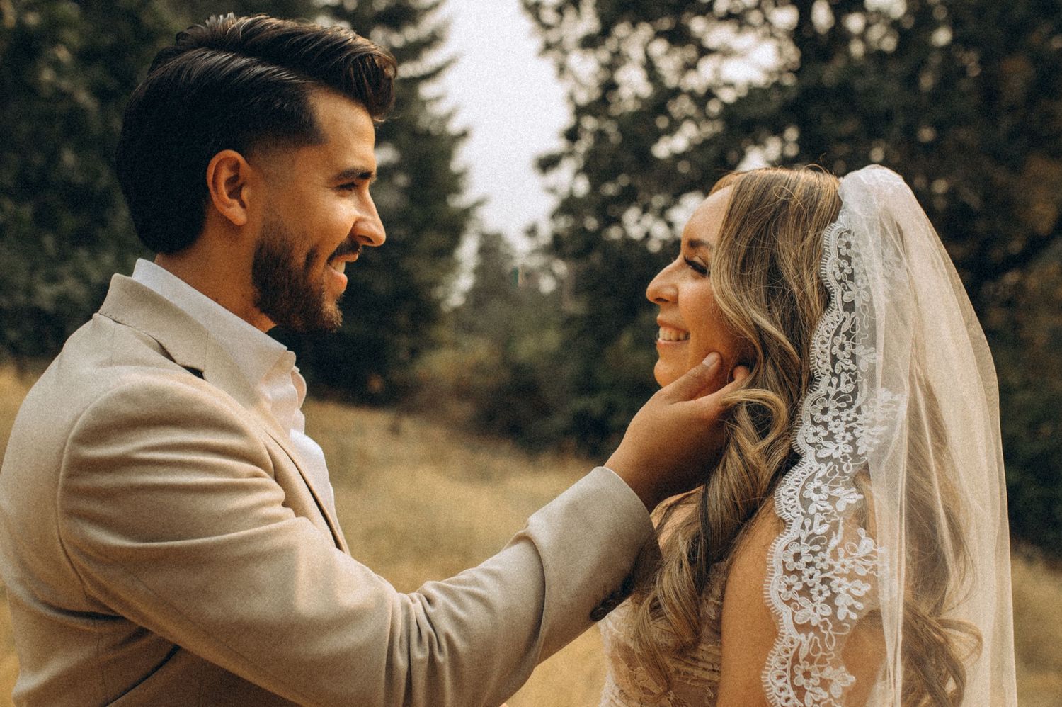 Tender moment between newlyweds in a rustic outdoor setting with bride wearing lace veil and groom in beige suit.