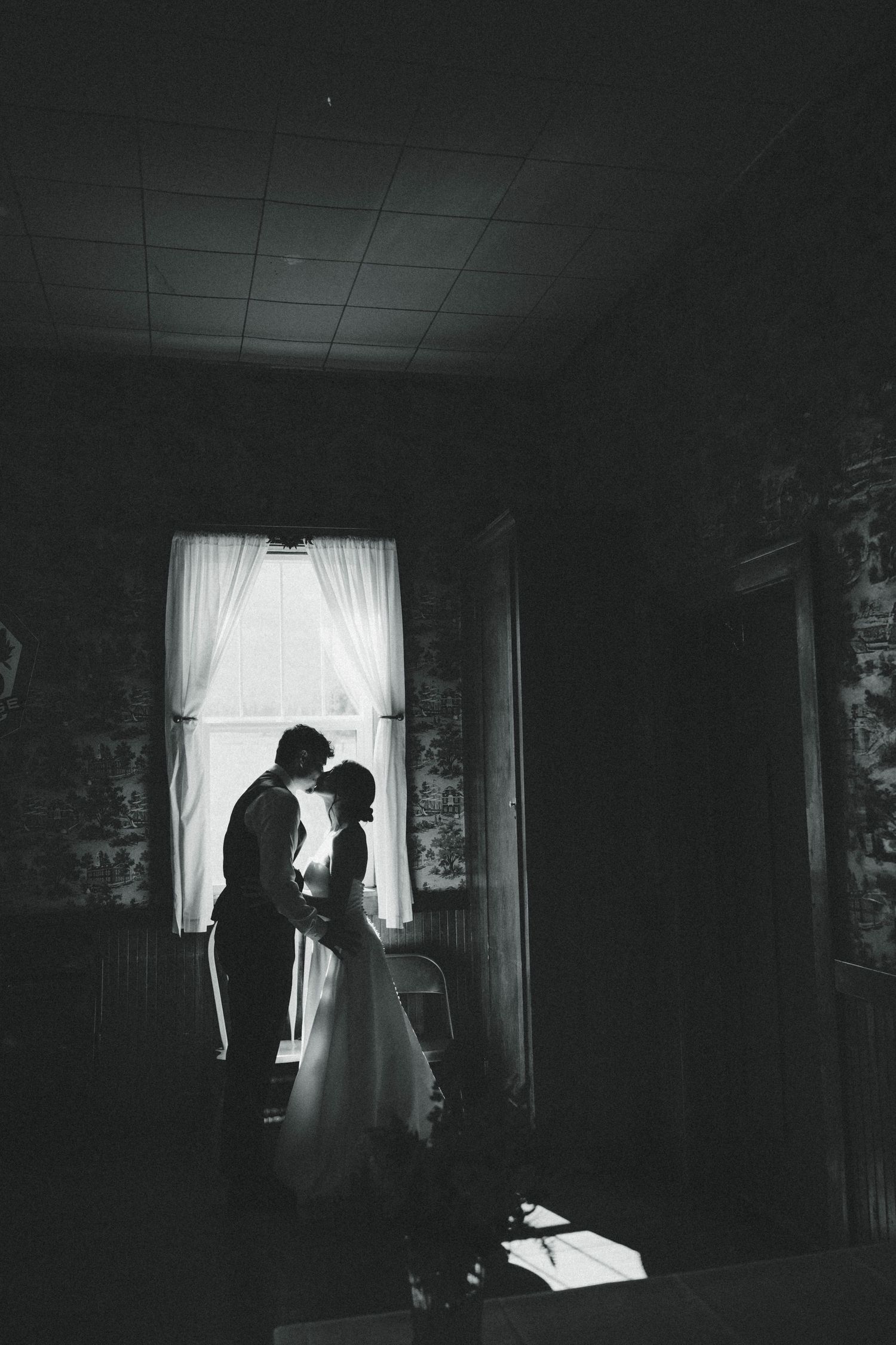 Black and white silhouette of a couple sharing a romantic moment by a window in a dimly lit room.